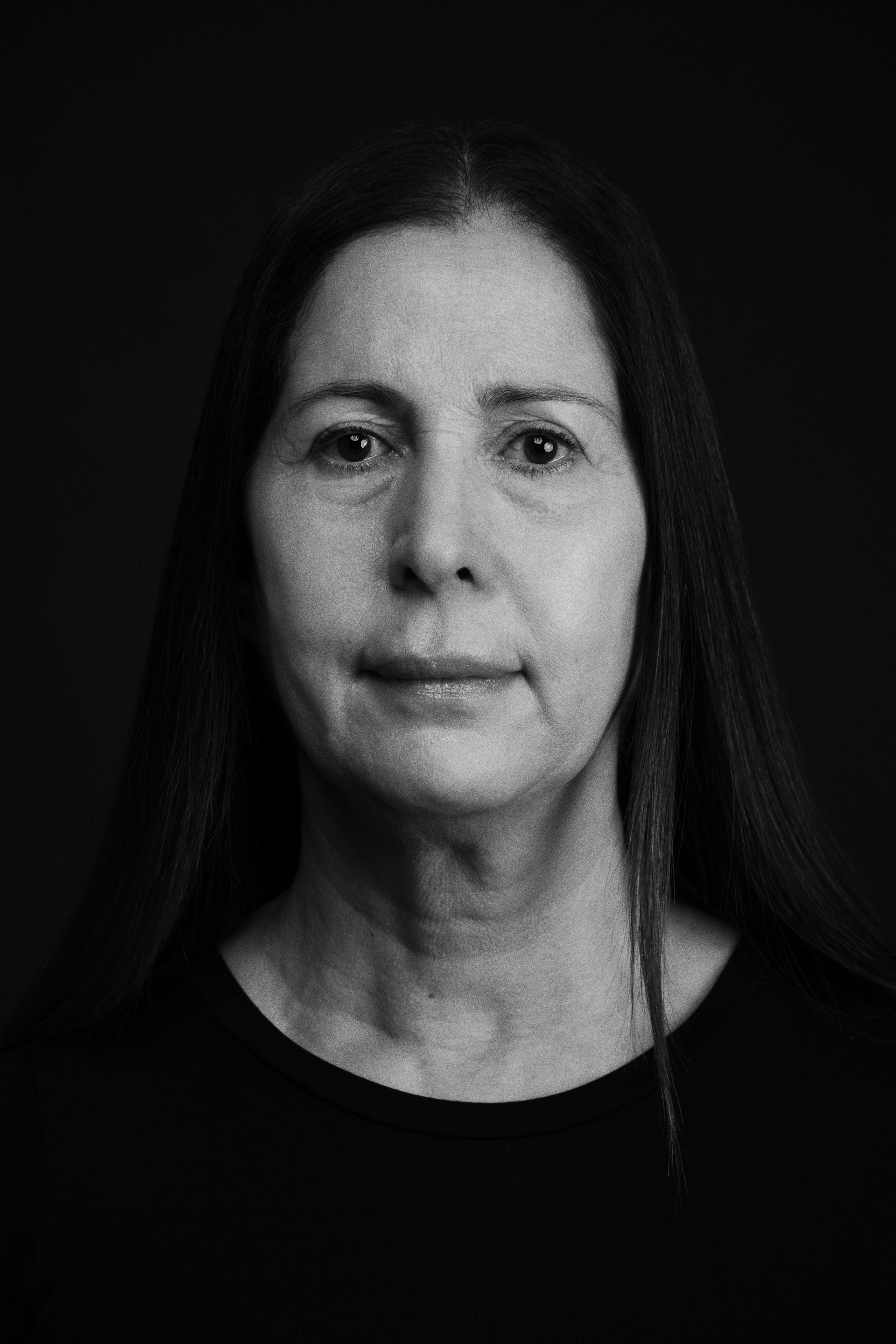 Black and white portrait of a middle-aged woman with long straight hair parted in the center, wearing a plain black crewneck top, looking directly at the camera with a calm, neutral expression against a solid dark background.