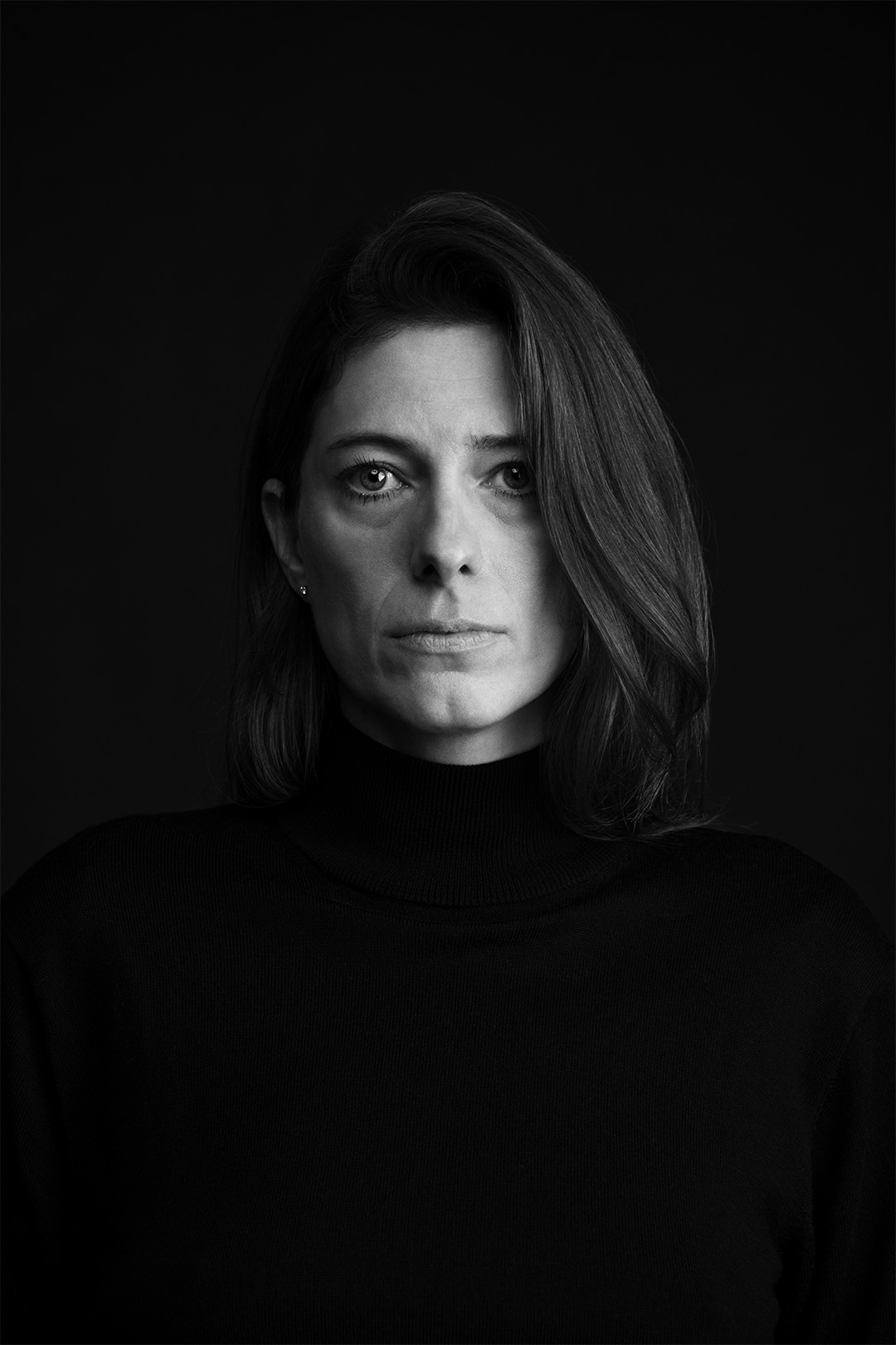 Black and white portrait of a woman with shoulder-length hair parted to the side, wearing a black turtleneck sweater, looking directly at the camera with a calm yet serious expression against a solid dark background.