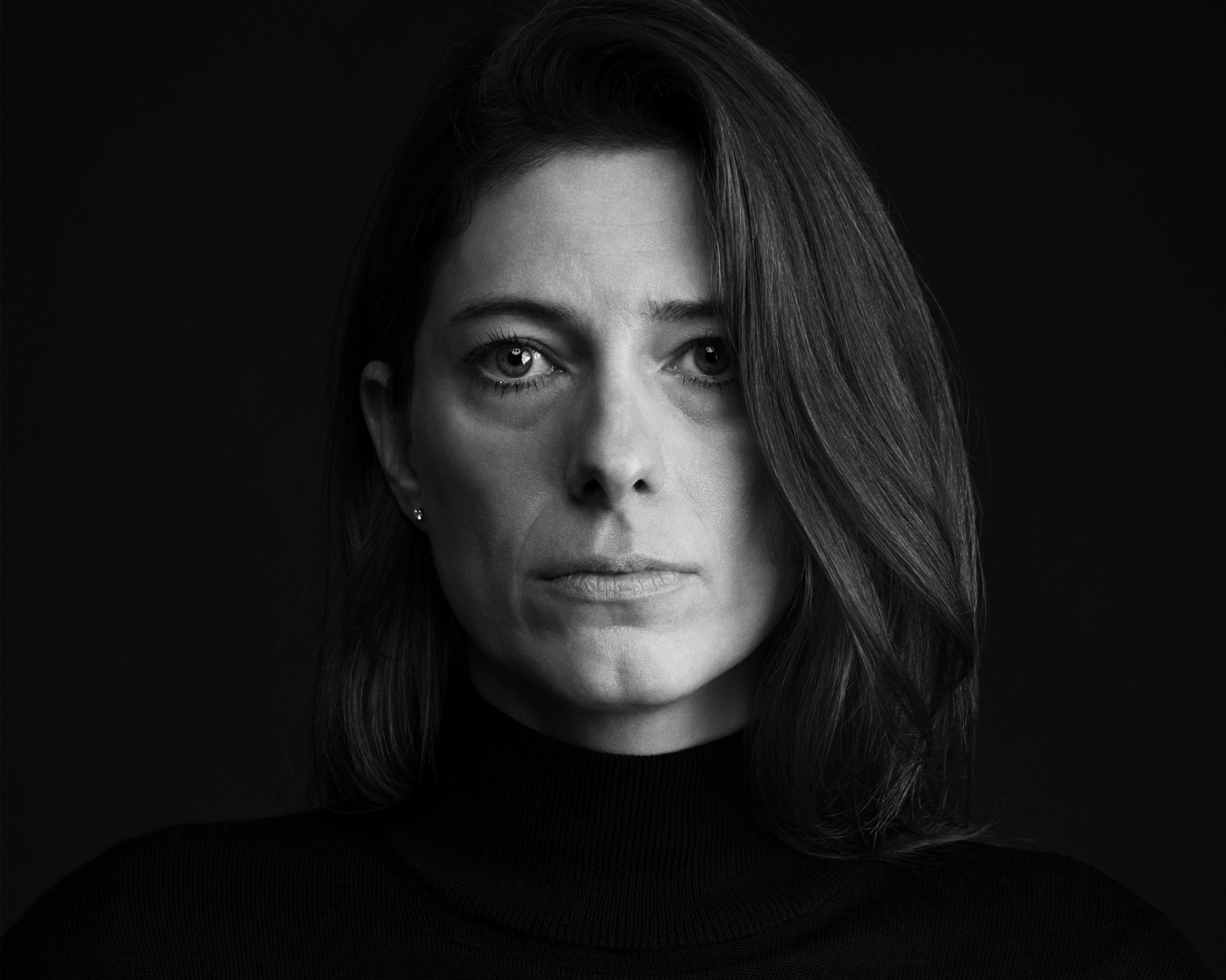 Black and white portrait of a woman with shoulder-length hair parted to the side, wearing a black turtleneck sweater, looking directly at the camera with a calm yet serious expression against a solid dark background.