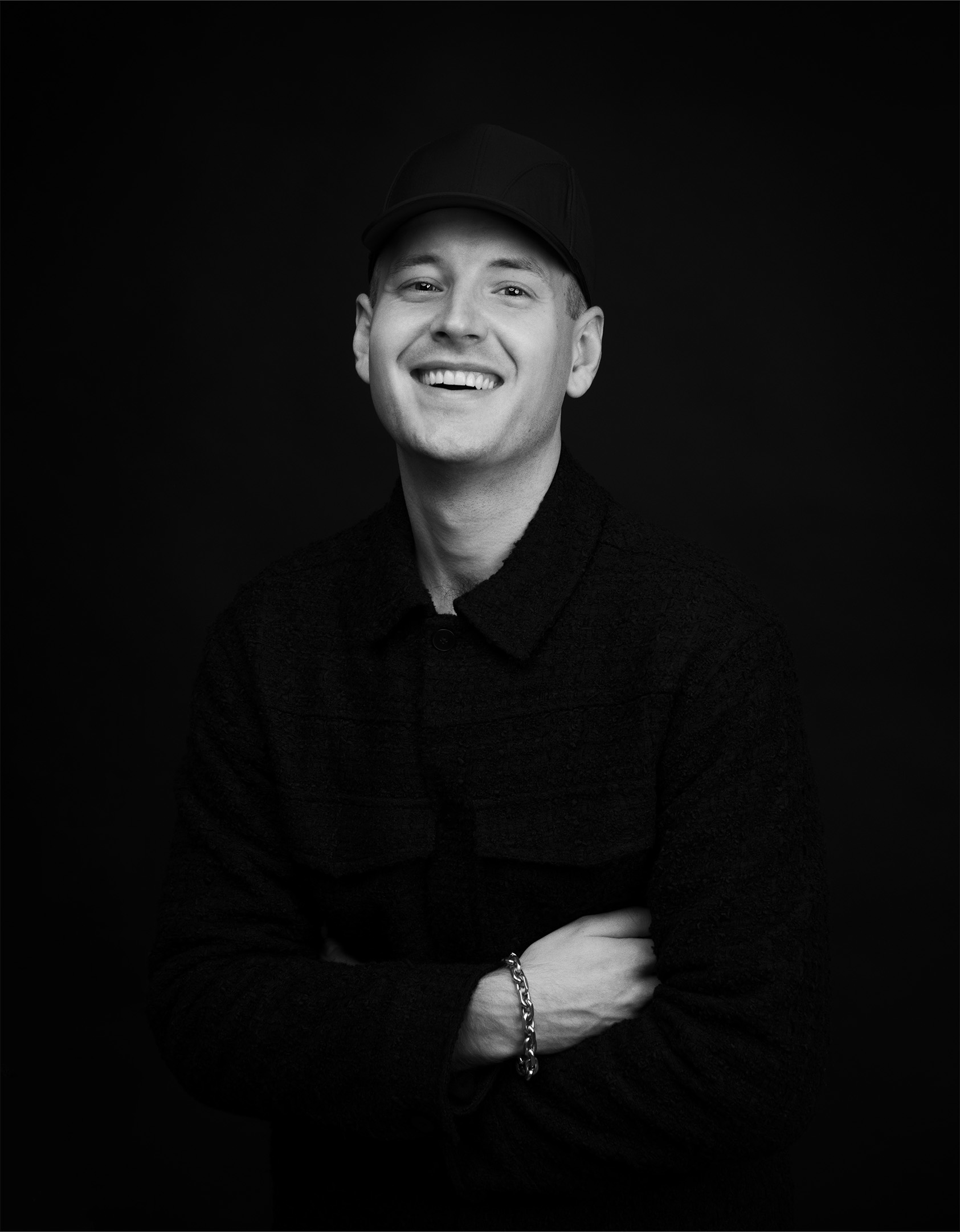 Black and white portrait of a smiling man wearing a dark baseball cap and textured jacket, posed against a plain dark background. He is looking directly at the camera with arms crossed, showing a chunky silver chain bracelet on his wrist. The high-contrast lighting emphasizes his facial expression and creates a clean, professional studio photography style.