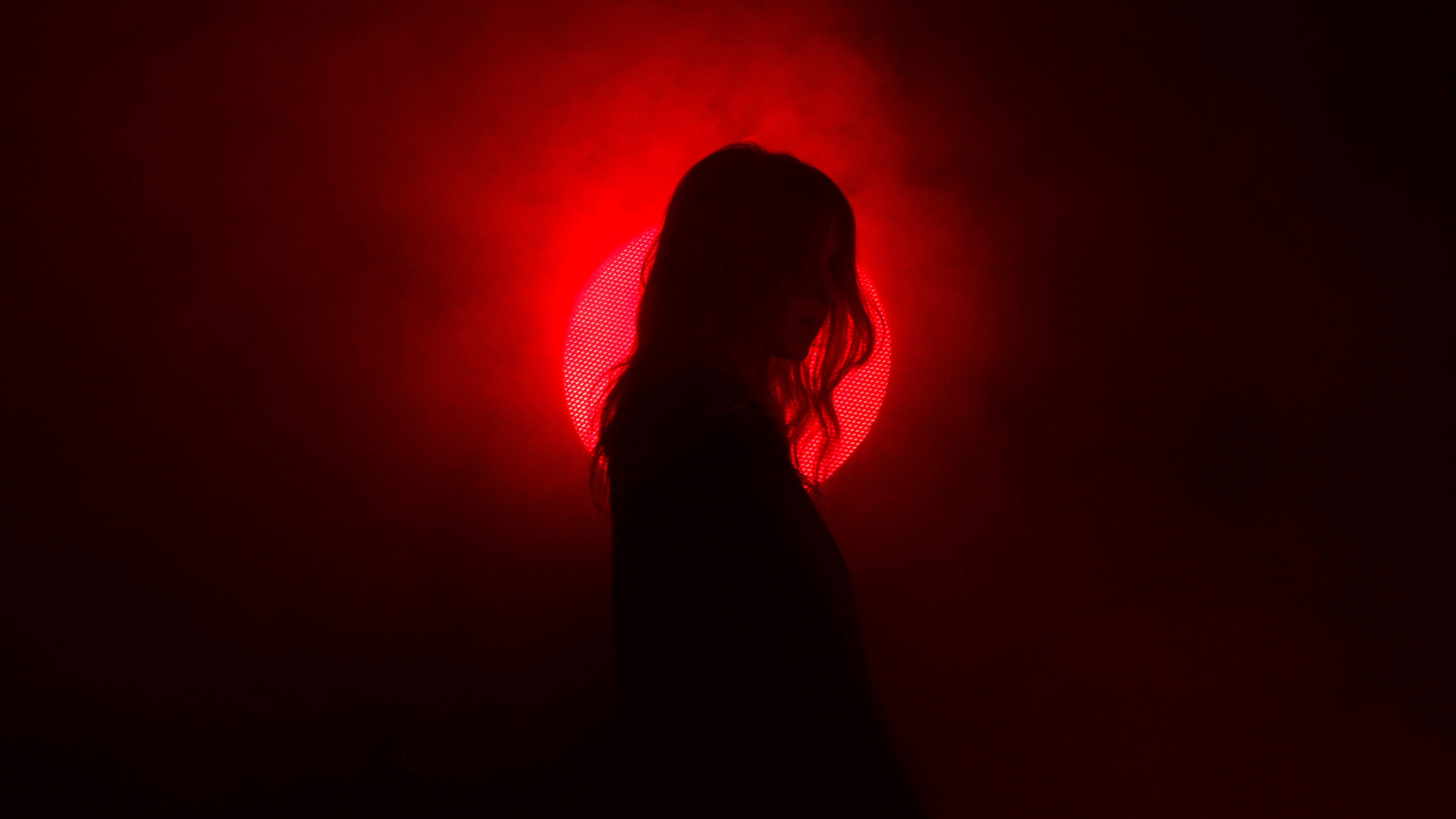 Silhouette of a person with long hair standing against a glowing red circular light, resembling a sun or moon, surrounded by smoky mist. The deep shadows conceal facial details, while the backlit red aura creates a dramatic and mysterious atmosphere. The composition emphasizes contrast, moody lighting, and a cinematic, almost surreal visual style.