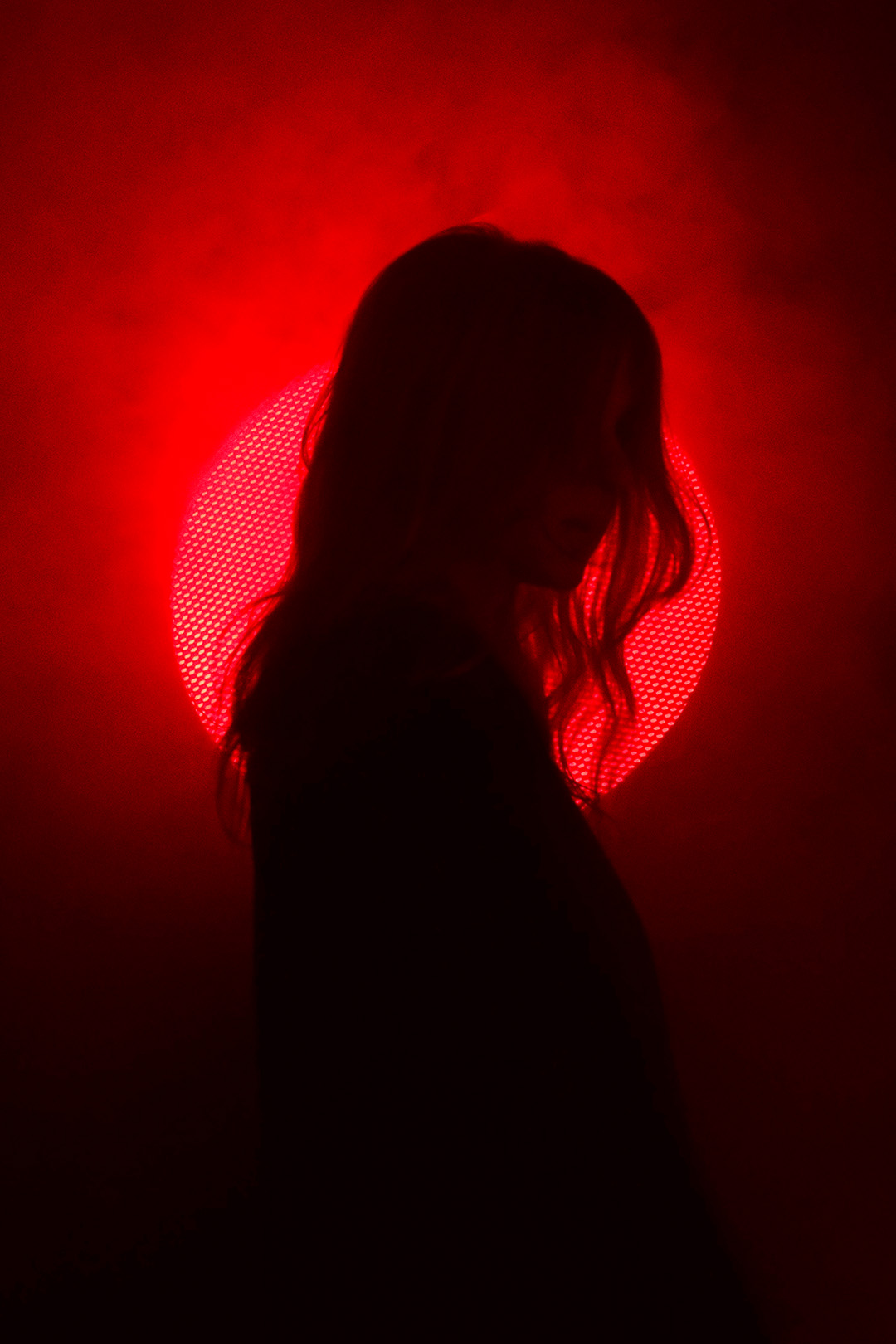 Silhouette of a person with long hair standing against a glowing red circular light, resembling a sun or moon, surrounded by smoky mist. The deep shadows conceal facial details, while the backlit red aura creates a dramatic and mysterious atmosphere. The composition emphasizes contrast, moody lighting, and a cinematic, almost surreal visual style.