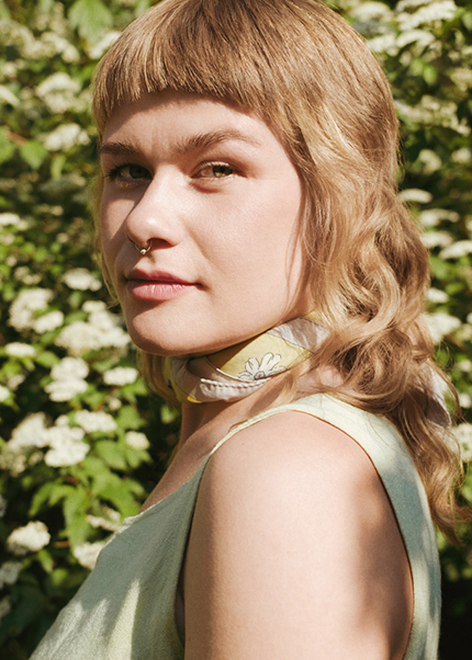 Close-up portrait of Katinka Askehave with light hair and scarf around her neck, standing among blooming white flowers and looking toward the camera.