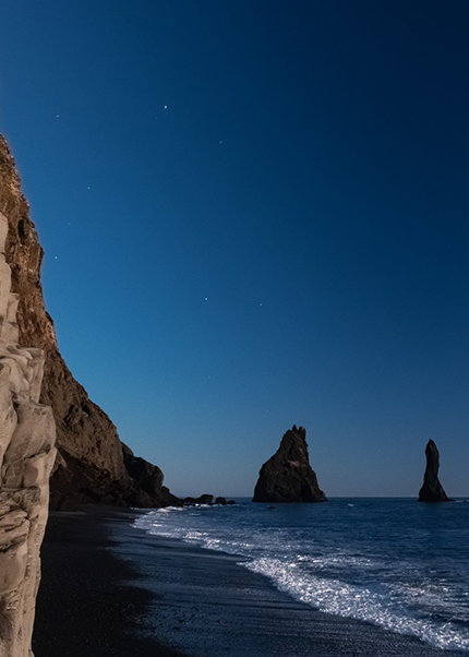 Color photo of basalt cliffs and sea stacks at Reynisfjara Beach in Iceland, with waves breaking on the black sand shore.