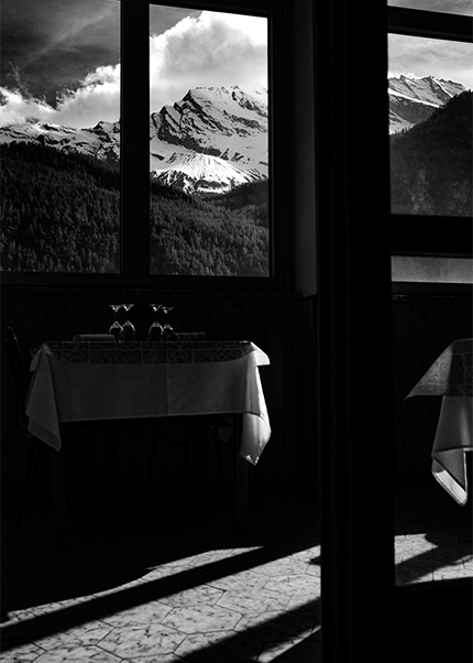 Black and white photo of Gran Paradiso mountains seen through a restaurant window with a table and shadows in the foreground.