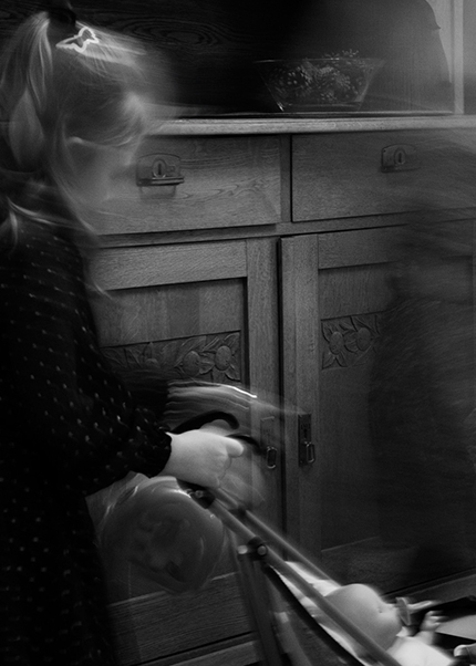 Black and white photo of a child pushing a doll’s pram with blurred ghostlike motion effect.
