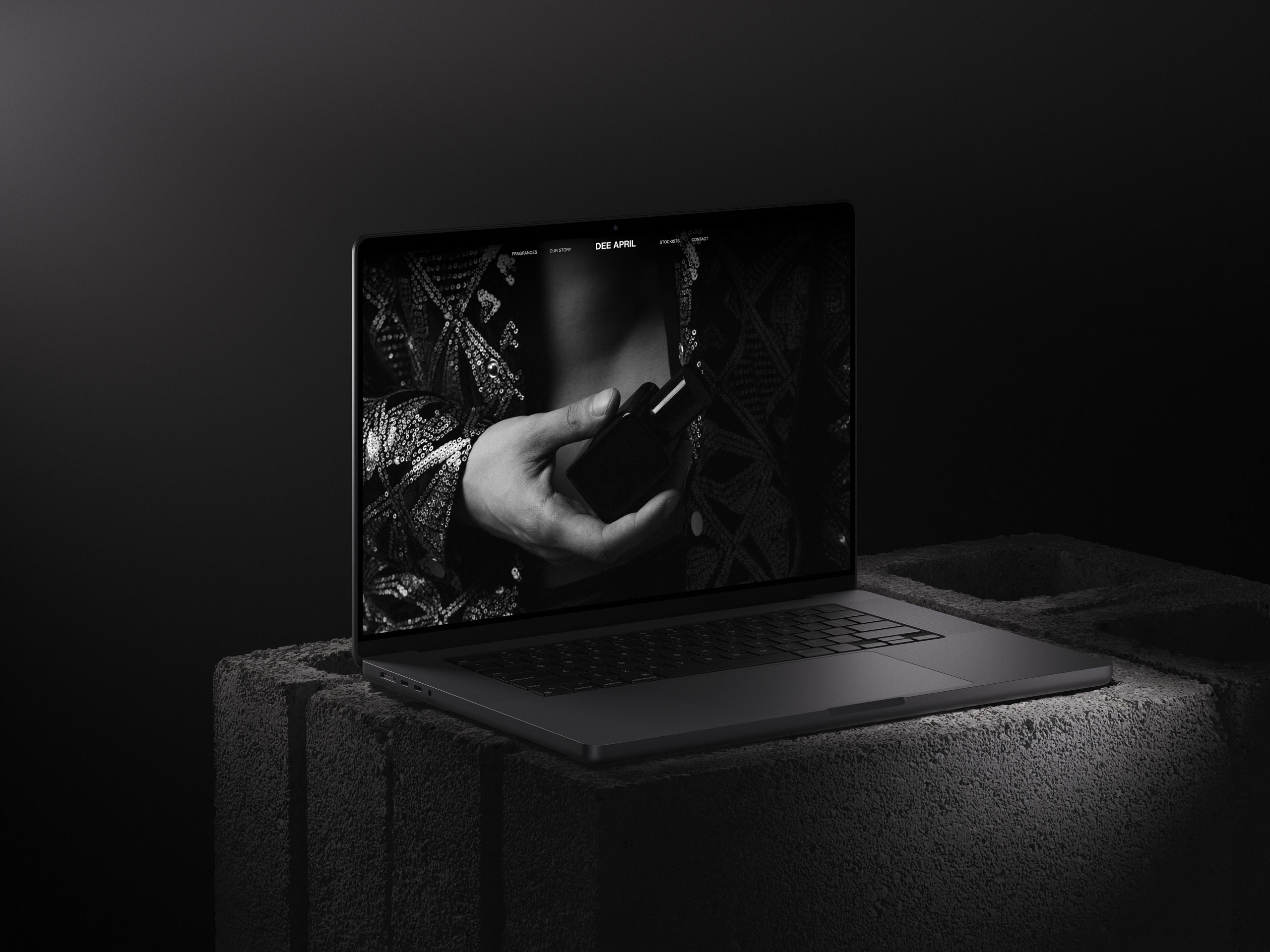 Laptop mockup showcasing DEE APRIL's black and white website design, featuring a hand holding a perfume bottle, placed on a textured concrete block.
