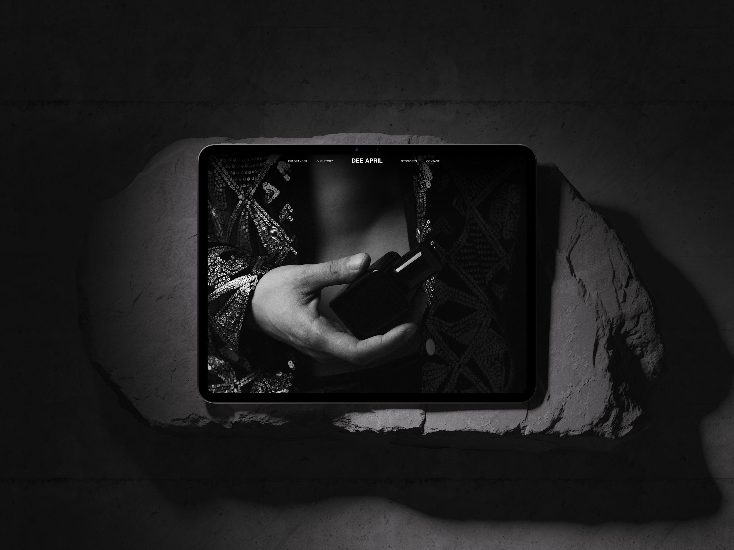 Tablet mockup presenting DEE APRIL's black and white fragrance brand website design, featuring a close-up hand holding a perfume bottle, placed on a rugged stone surface.