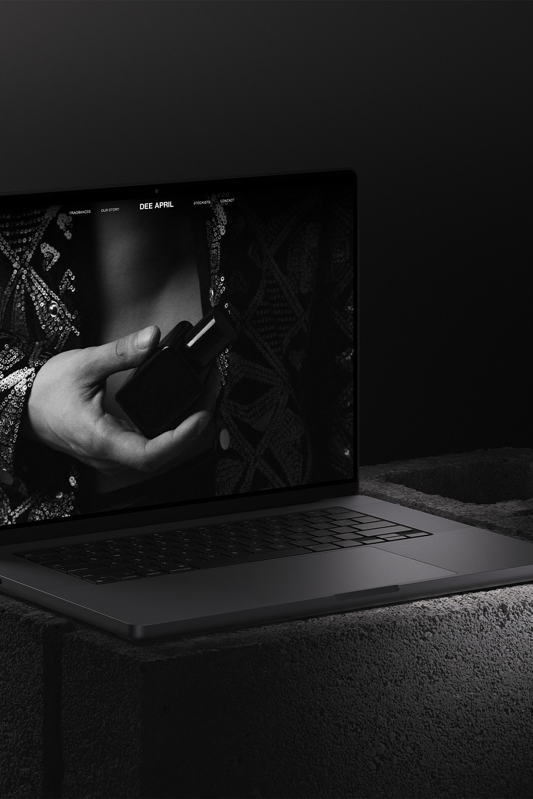 Laptop mockup showcasing DEE APRIL's black and white website design, featuring a hand holding a perfume bottle, placed on a textured concrete block.