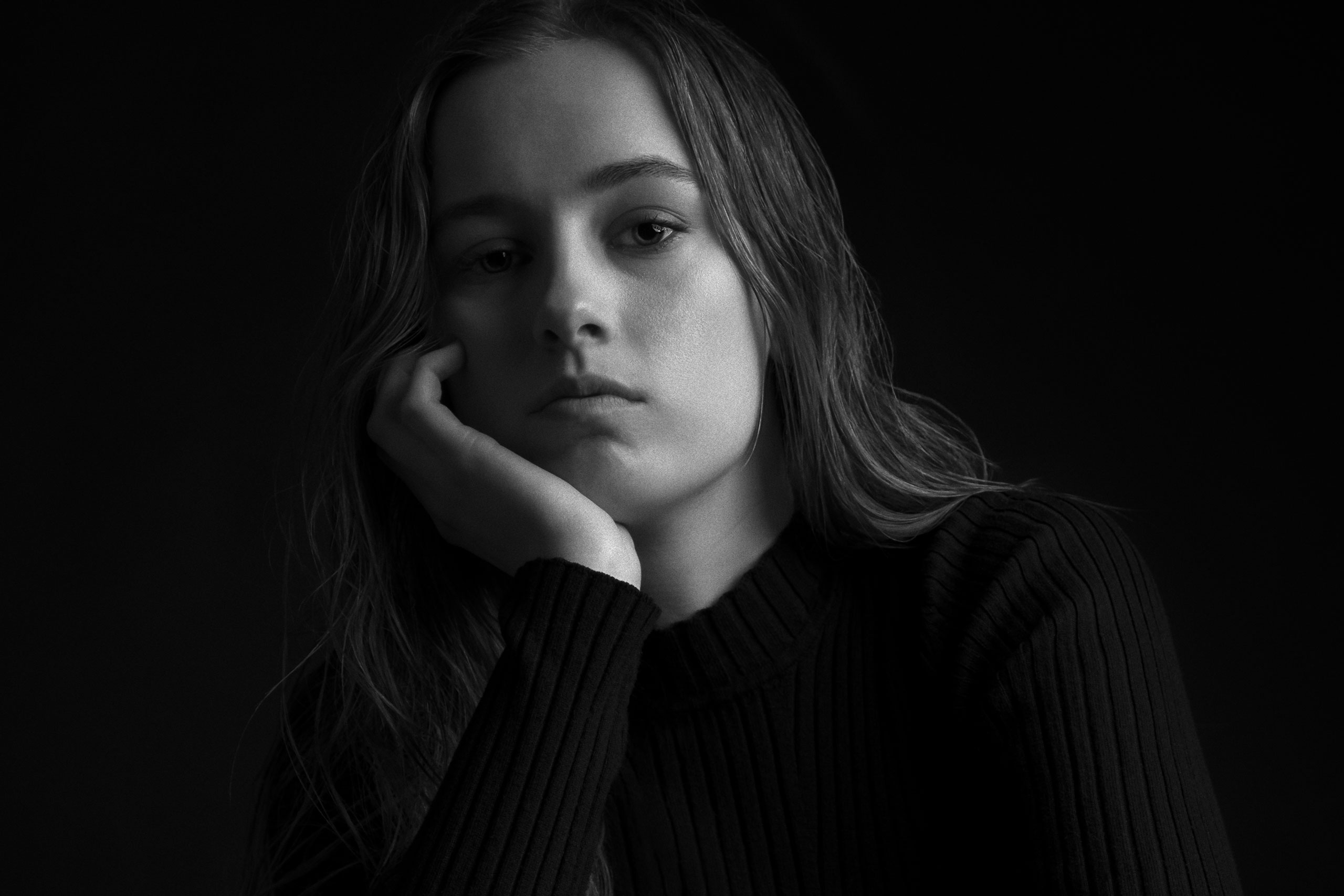 Black and white portrait of Maria-May Backhaus Brown in a ribbed dark sweater, resting her face on her hand, photographed against a dark background.