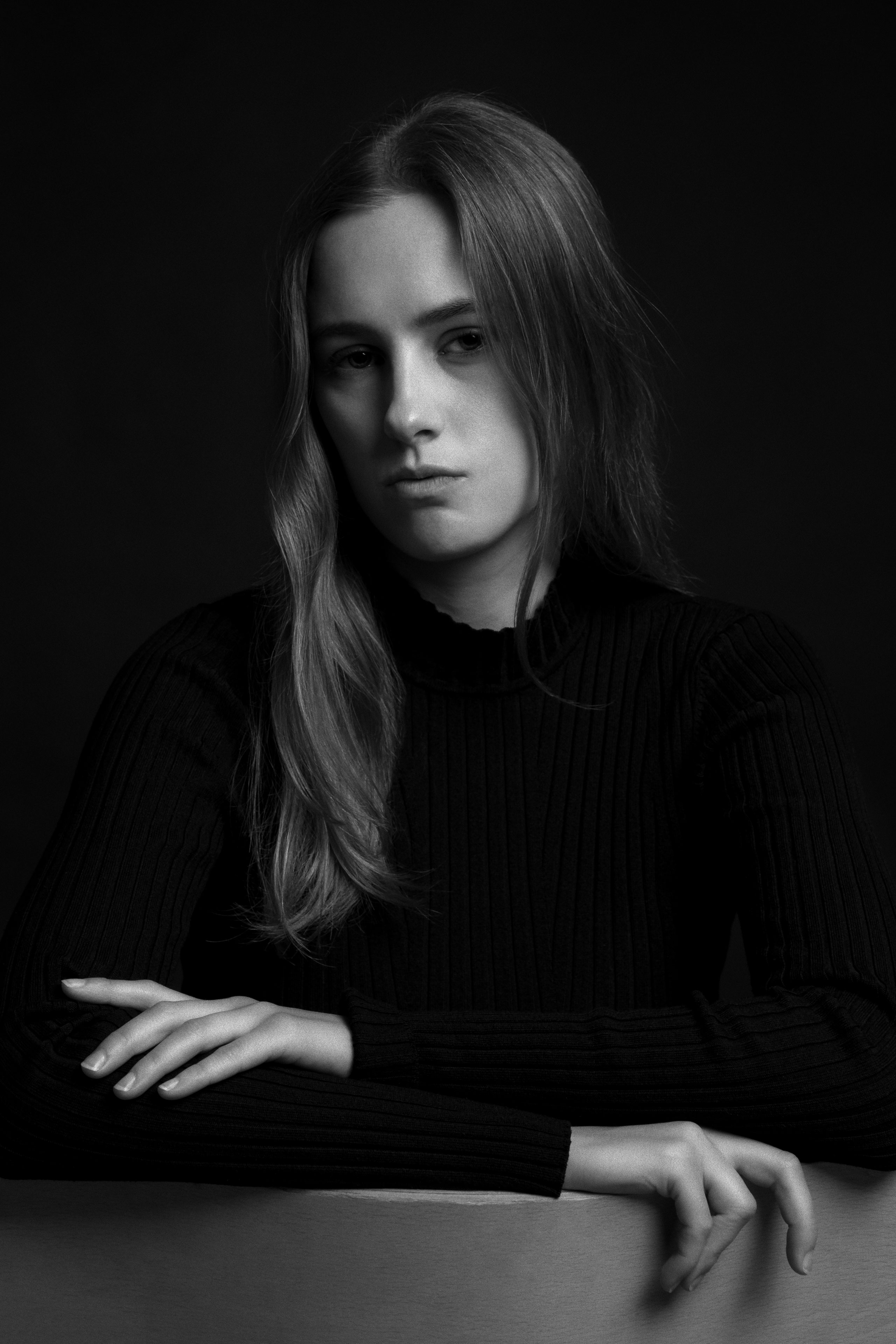 Black and white portrait of Maria-May Backhaus Brown in a ribbed dark sweater, seated with her arms resting on a surface, looking directly at the camera against a dark background.