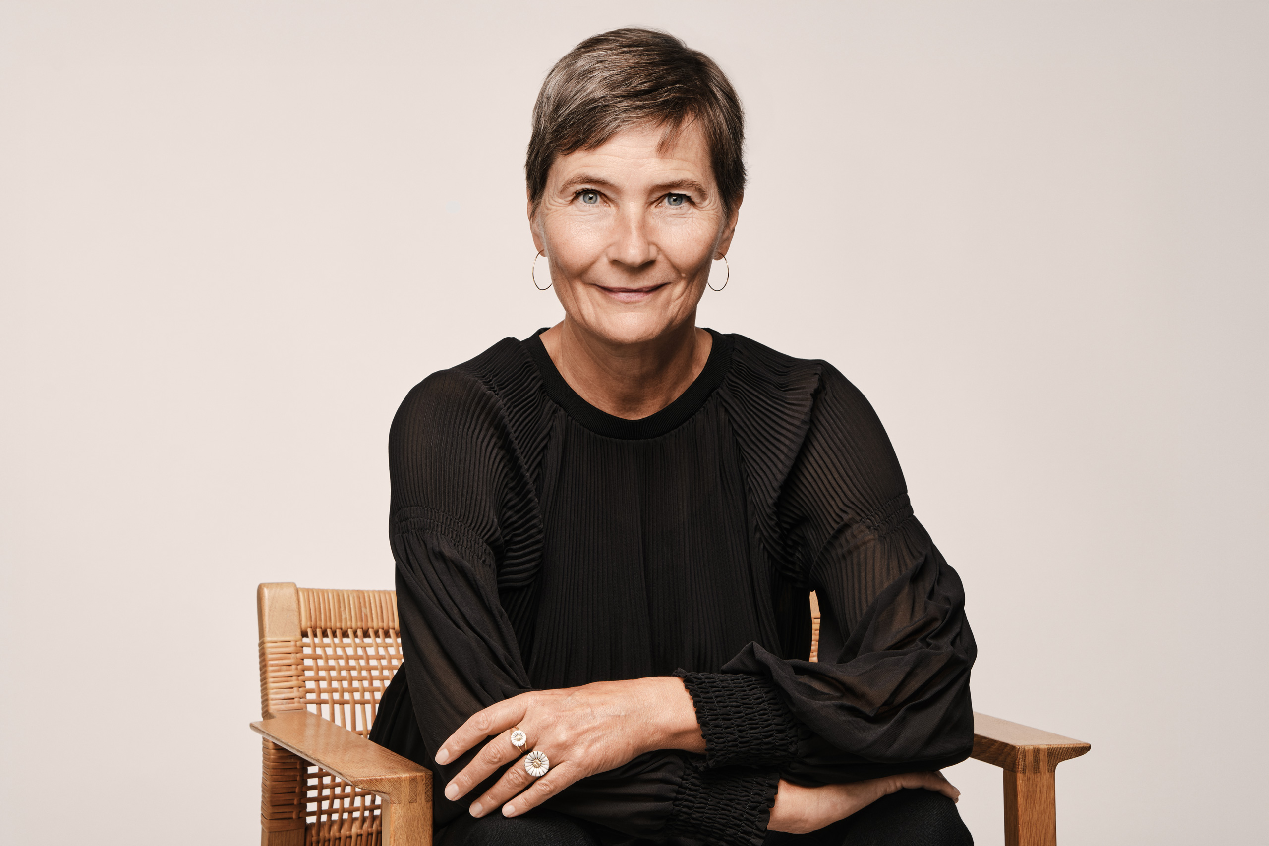 A professional studio portrait of Lene Dammand Lund, Rector of the Royal Danish Academy, seated in a wooden chair against a neutral background, wearing a black blouse.