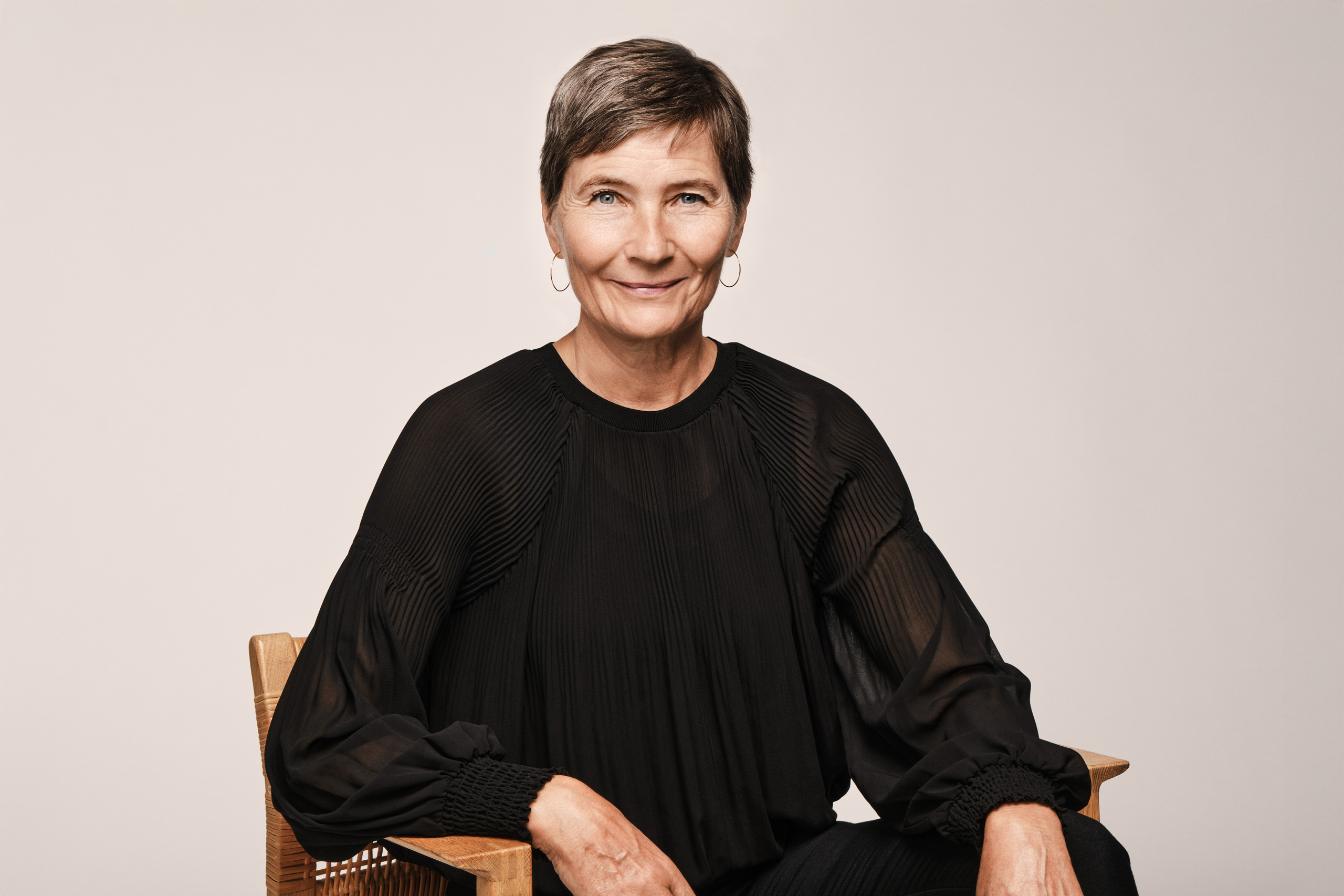 A professional studio portrait of Lene Dammand Lund, Rector of the Royal Danish Academy, seated in a wooden chair against a neutral background, wearing a black blouse.