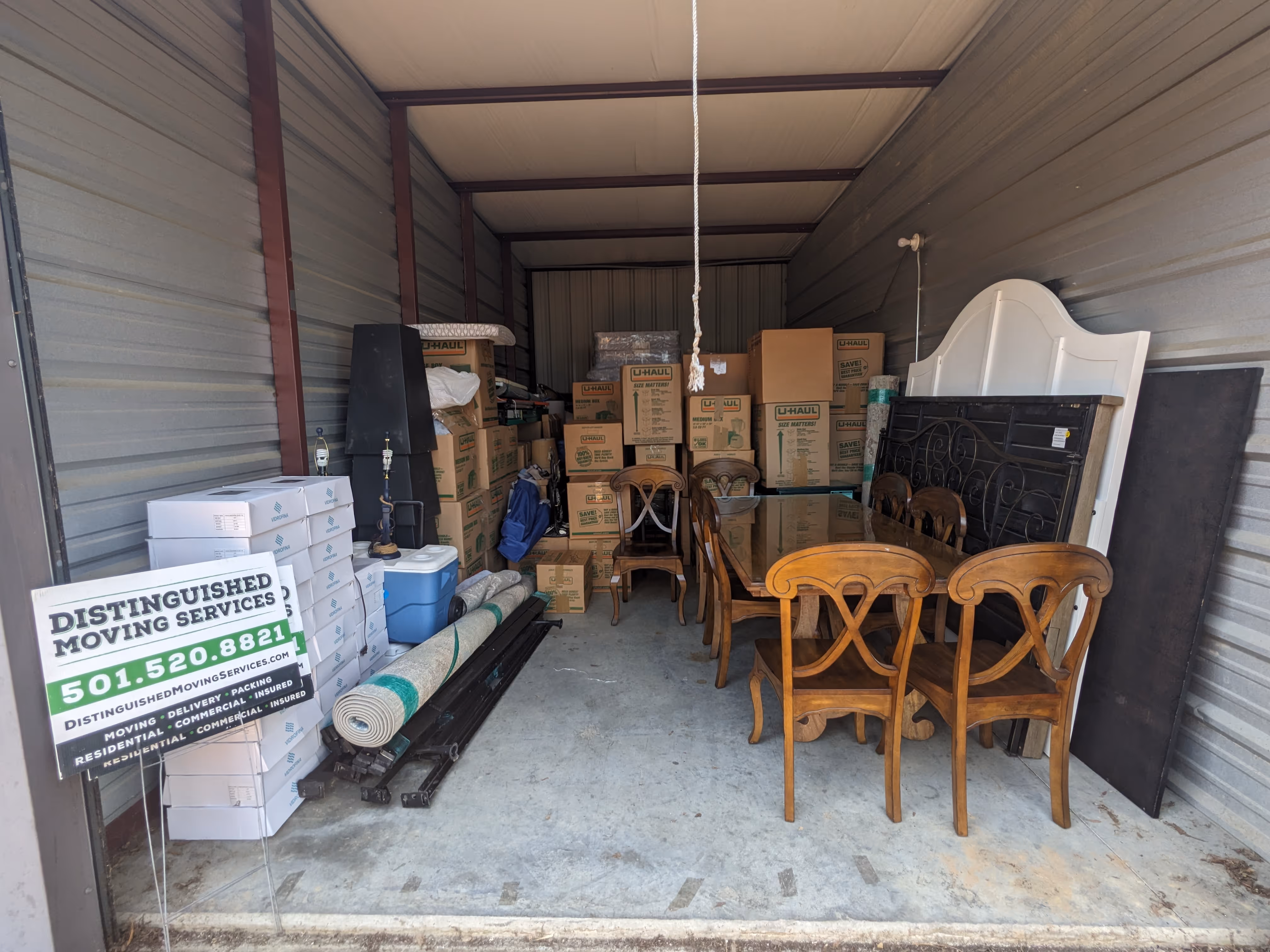 Storage unit filled with stacked U-Haul moving boxes, a wooden dining table with chairs, rolled-up carpet, and various household items.