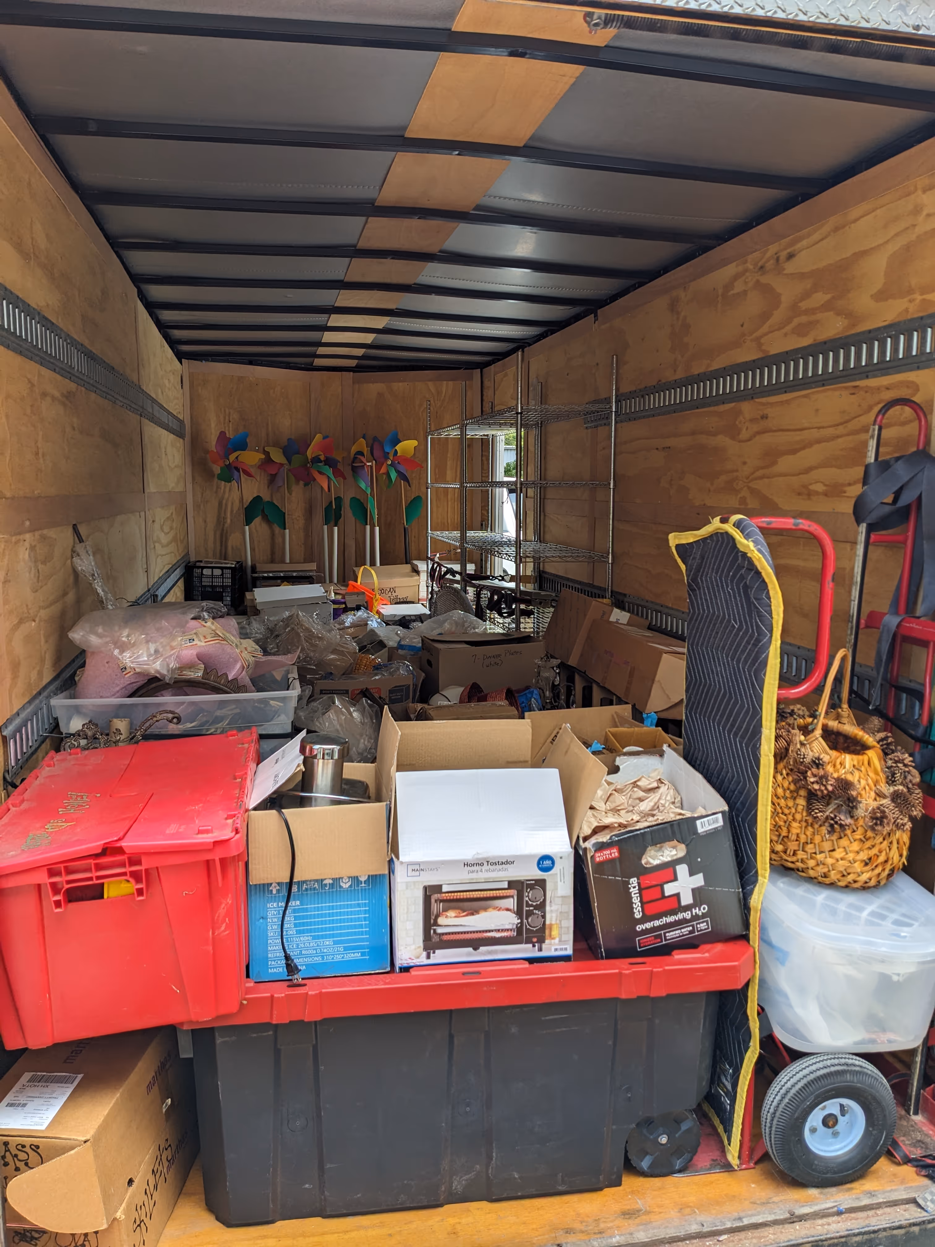 Back of a moving truck filled with various boxes, plastic bins, a metal shelving unit, colorful pinwheels, a woven basket, and a dolly.