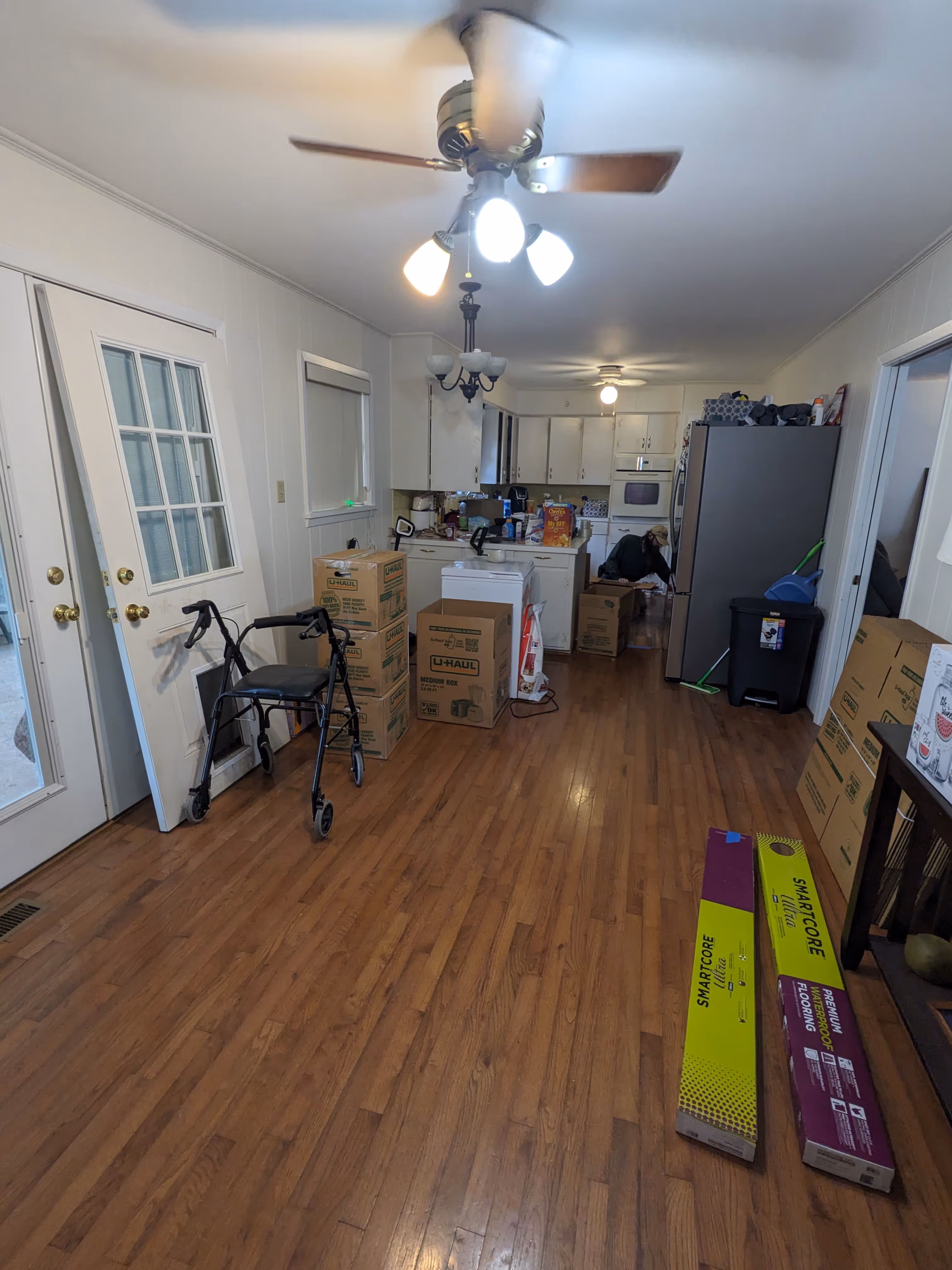 Kitchen and dining area with hardwood floors, cardboard moving boxes, a walker, and a person sitting near the back, organizing items.
