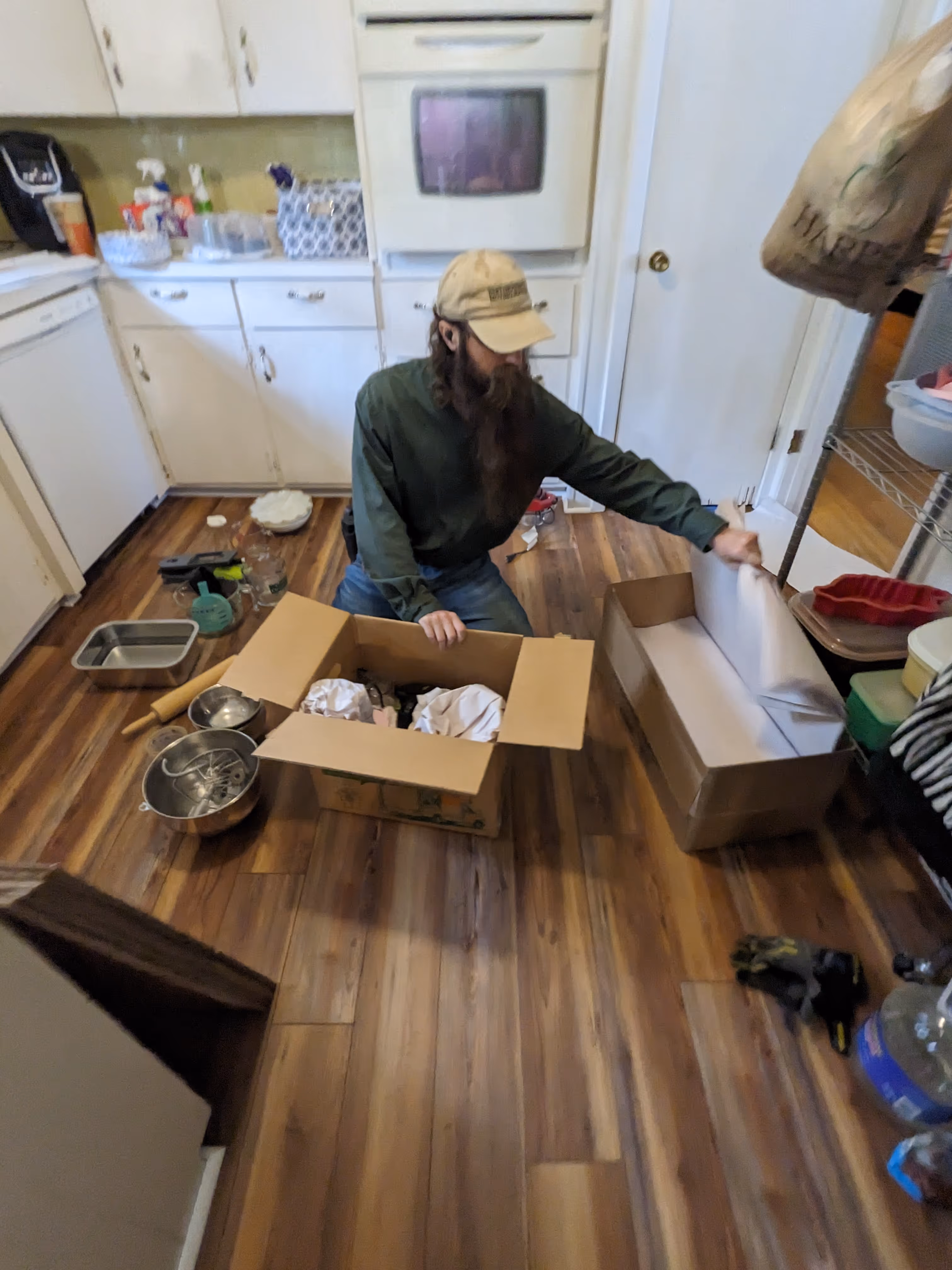 Man with a long beard and cap packing or unpacking boxes in a kitchen with wooden floors.