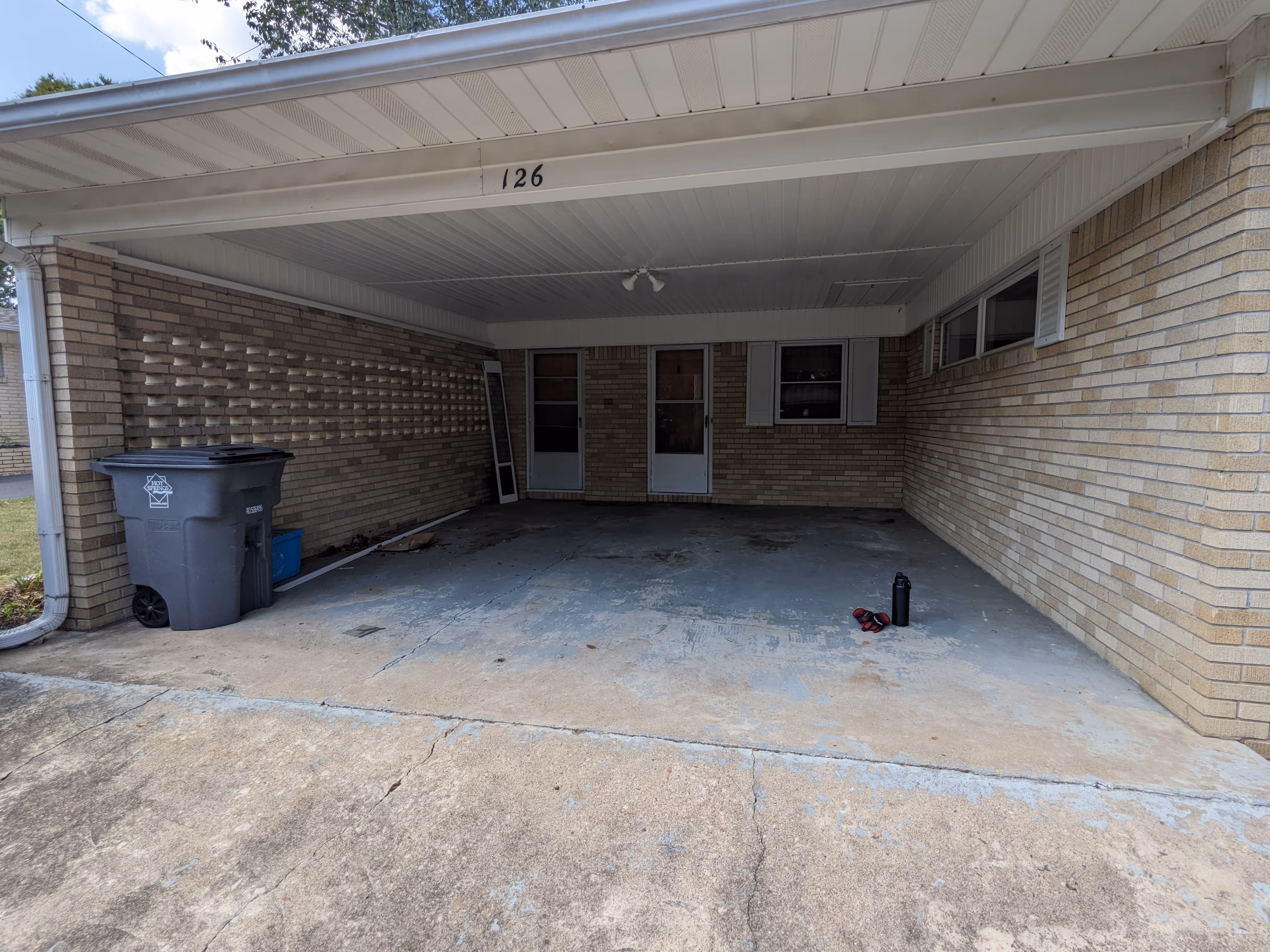 Covered carport with beige brick walls, concrete floor, trash bin, a water bottle, and gloves on the ground.