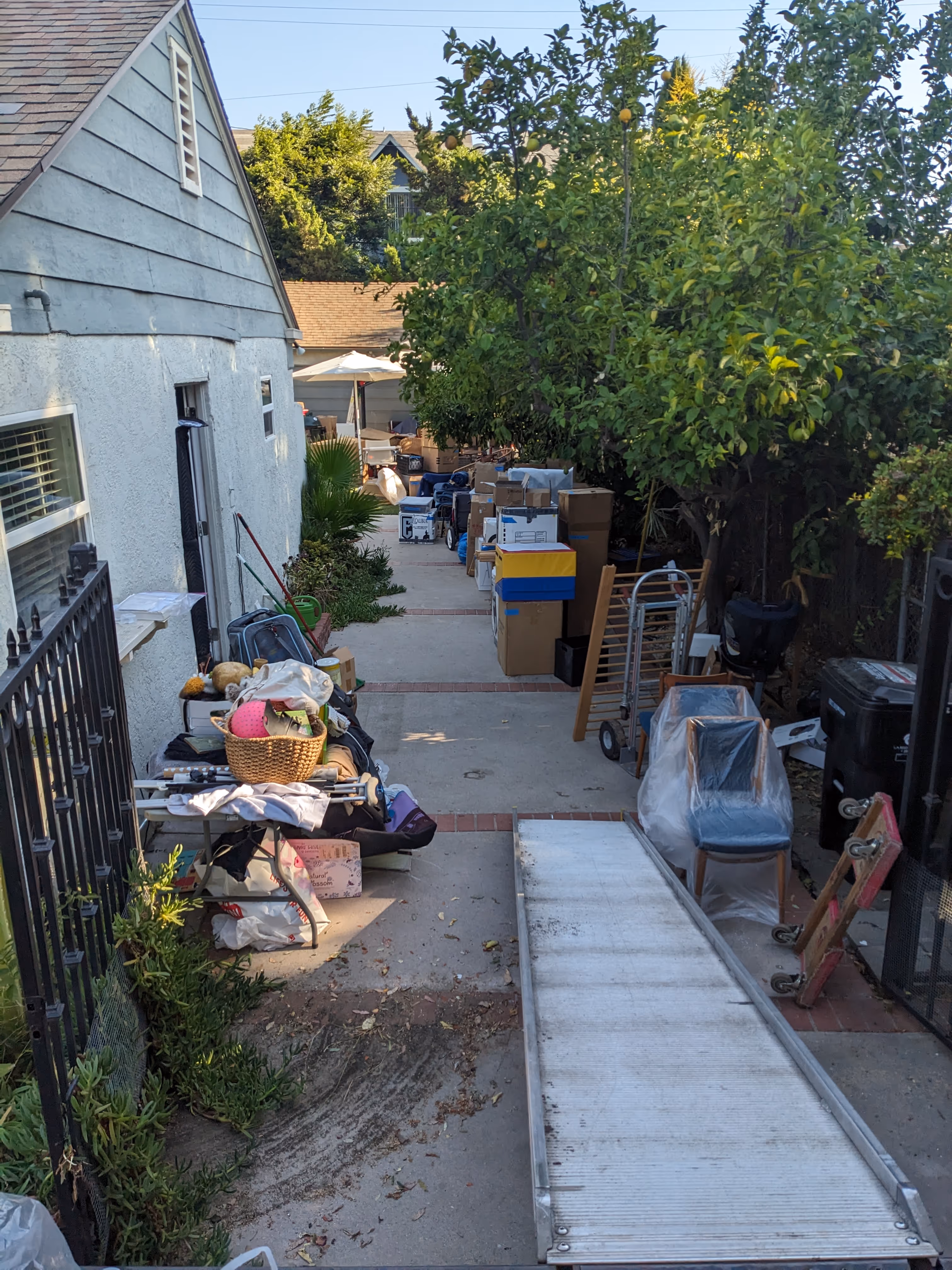 Narrow outdoor walkway with various packed boxes, furniture covered with plastic, a metal ramp, and household items alongside a white house and trees.