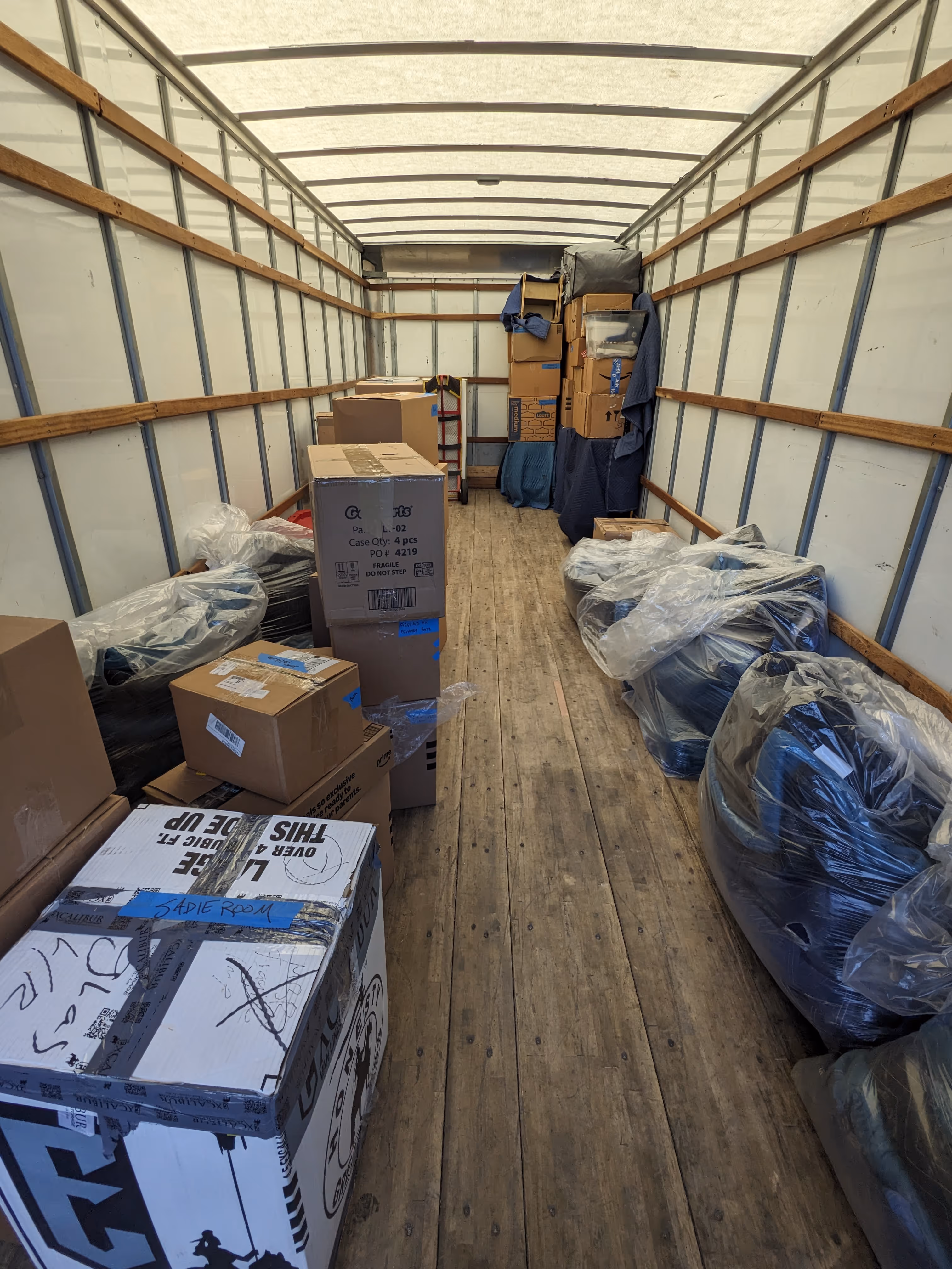Interior view of a moving truck with stacked cardboard boxes and plastic-covered furniture along the sides.