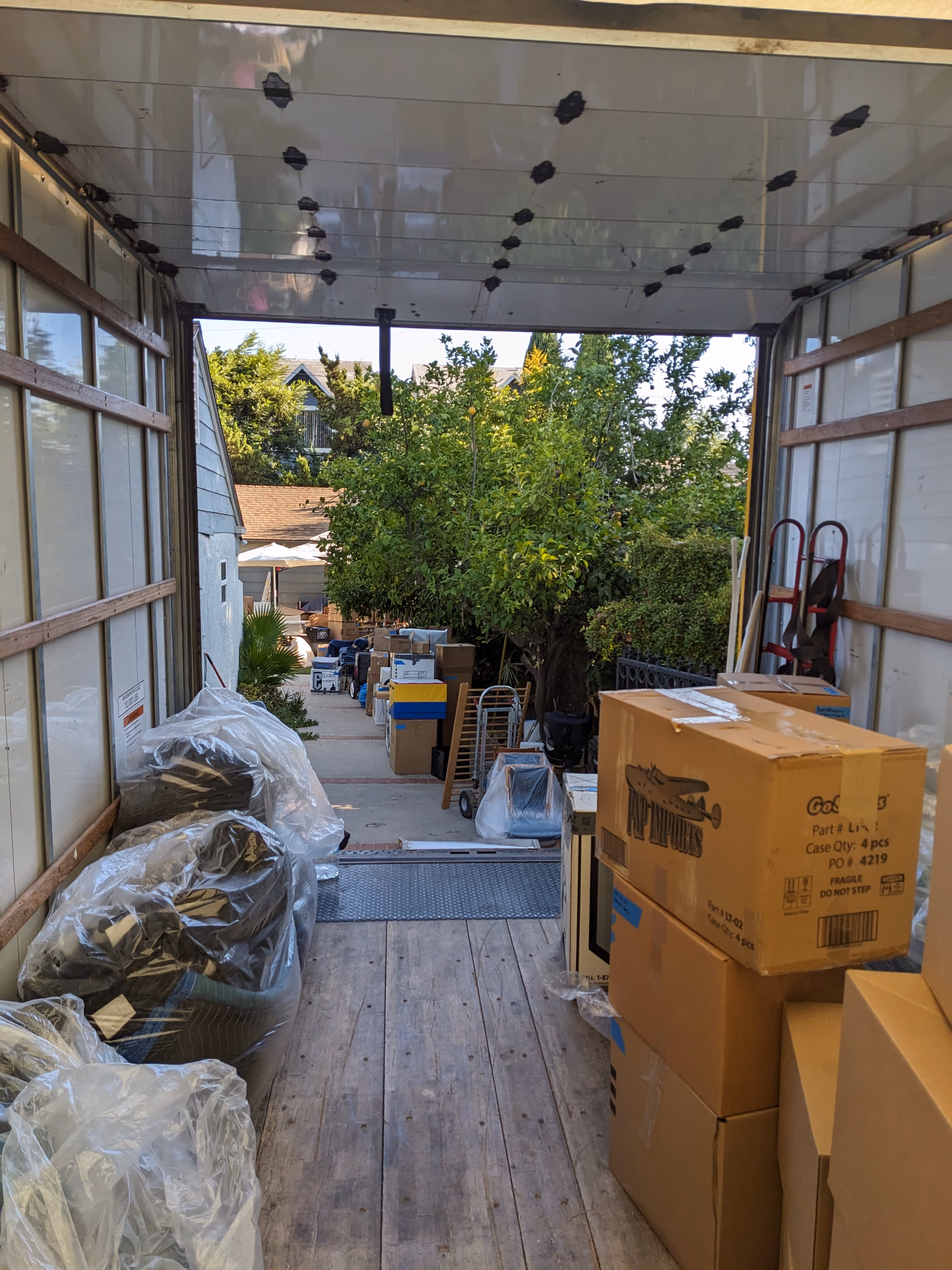 View from inside a moving truck showing packed cardboard boxes and furniture wrapped in plastic, with a sidewalk and trees outside.