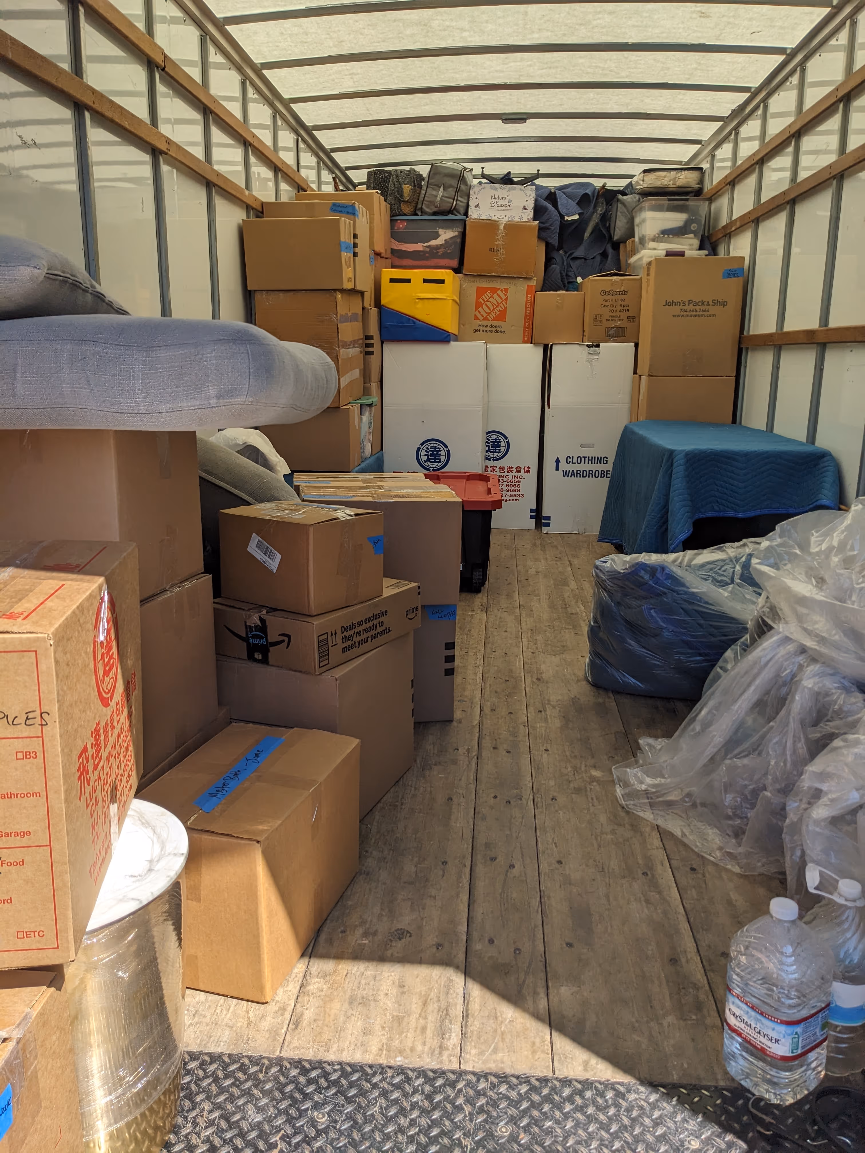 Interior of a moving truck loaded with cardboard boxes, plastic-wrapped items, and furniture covered with blankets.