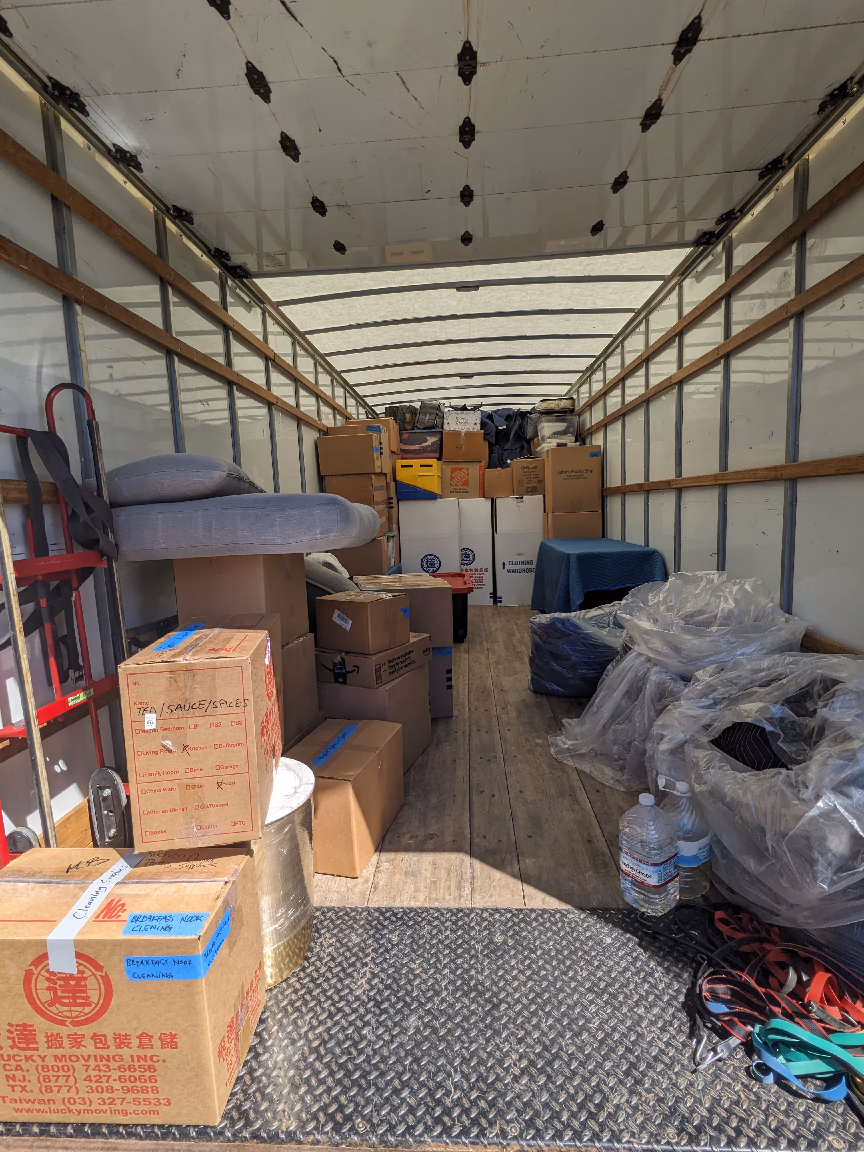 Interior of a moving truck loaded with packed cardboard boxes, wrapped furniture, plastic water bottles, moving straps, and a dolly.