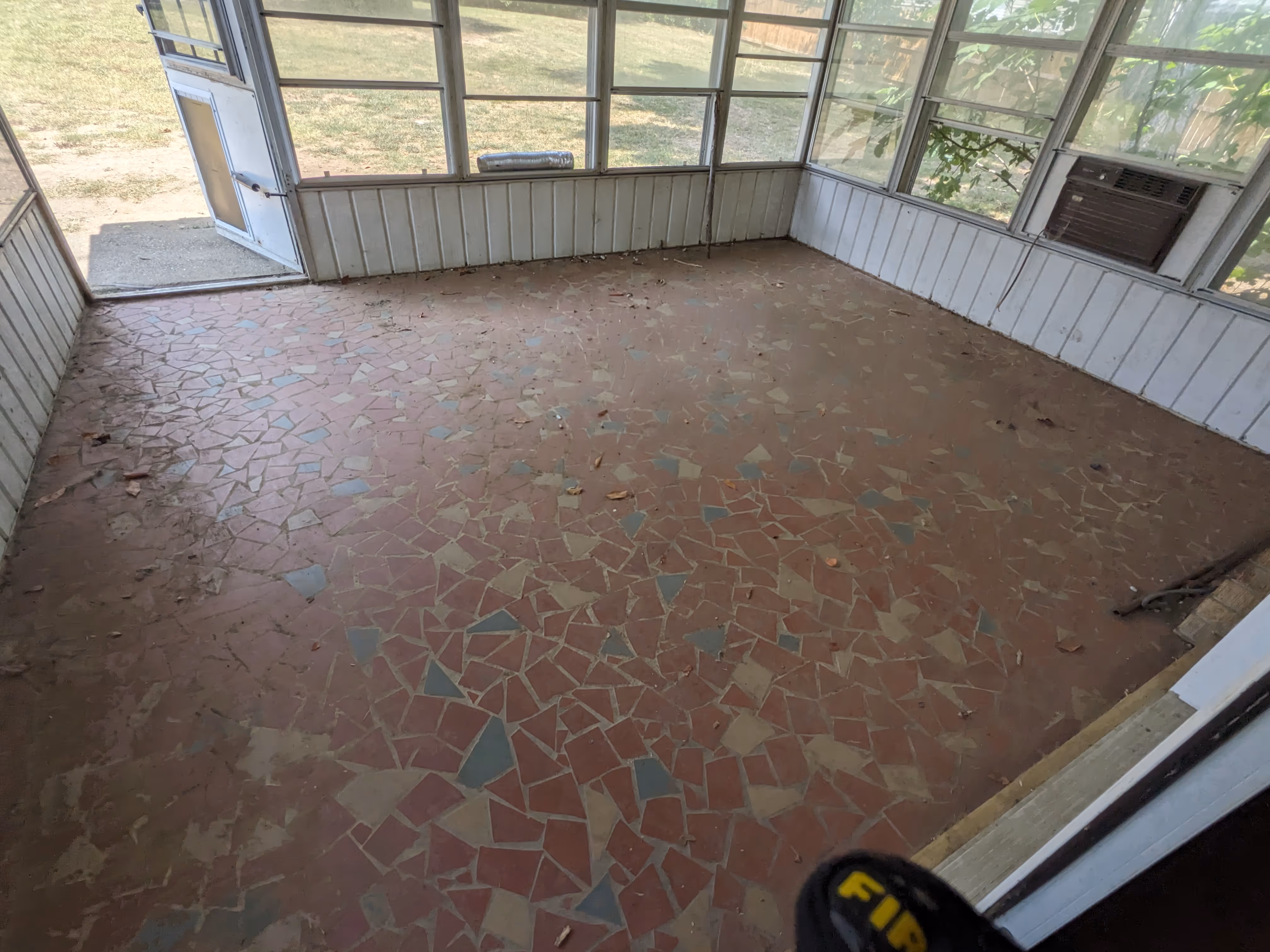 Empty sunroom with mosaic tile floor, white paneling, multiple windows, and an open door leading outside.