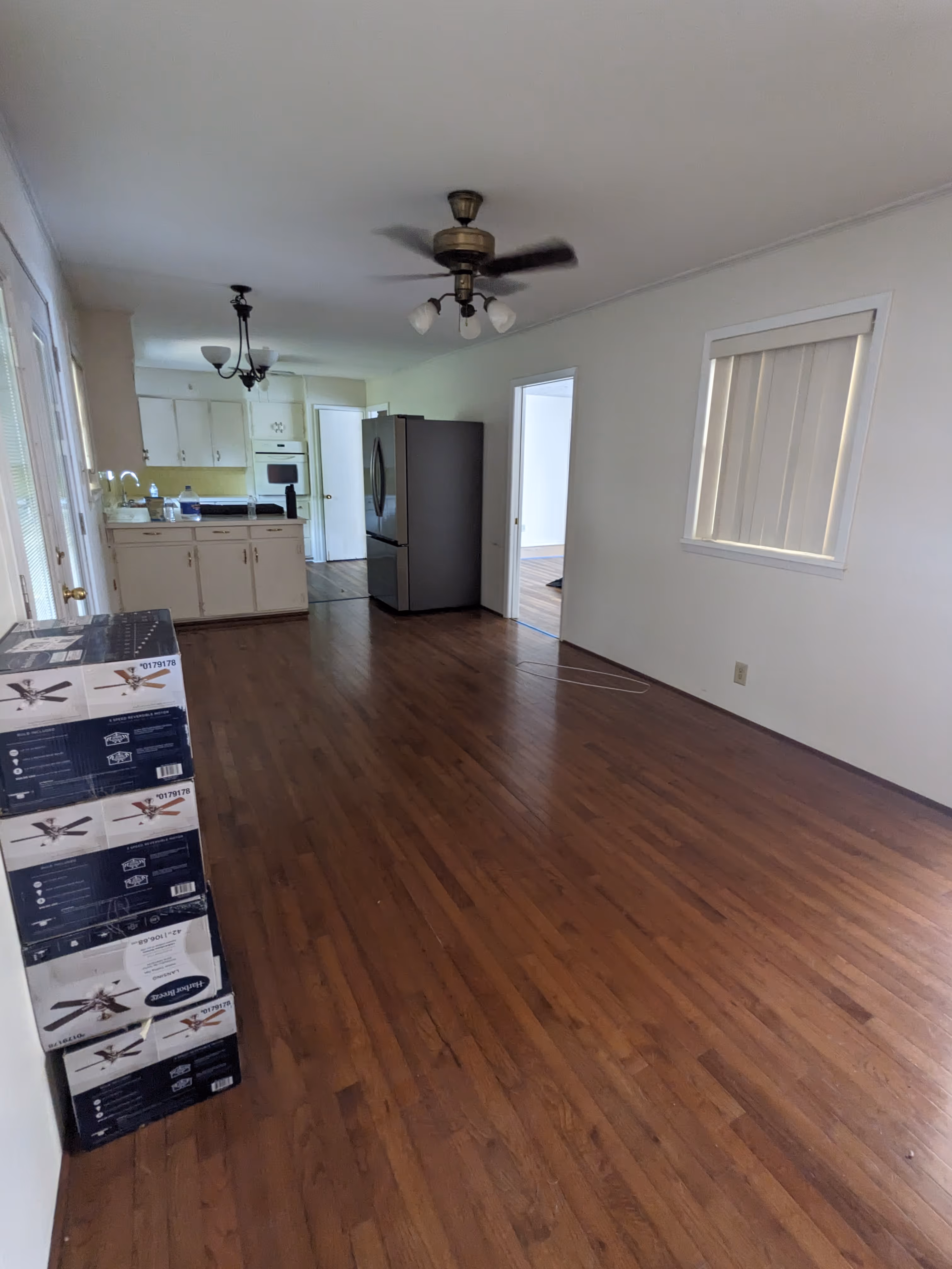Empty room with wooden floor, white walls, ceiling fan in motion, modern kitchen in the background, and stacked ceiling fan boxes on the left.