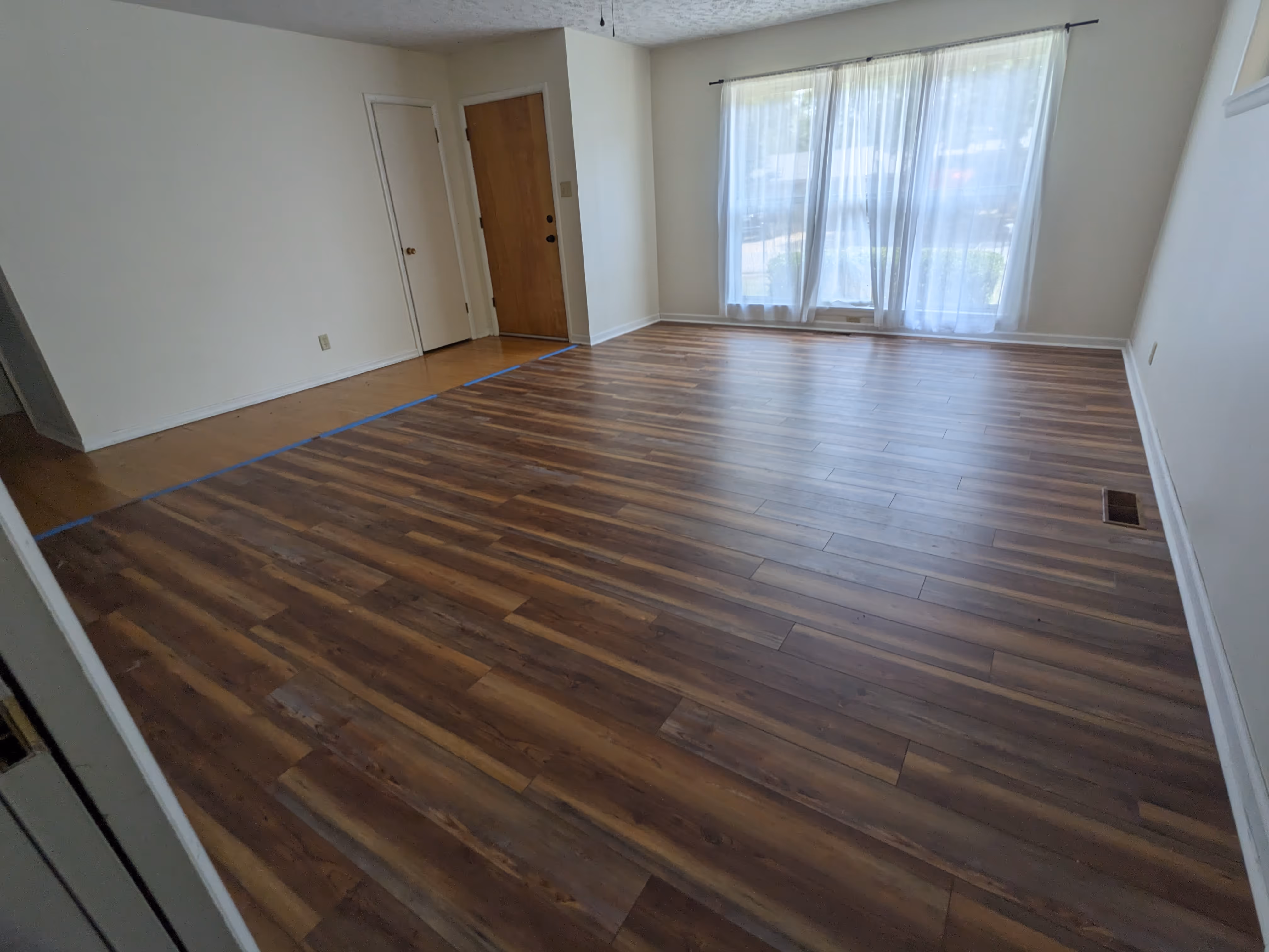 Empty room with new wood laminate flooring and large window with sheer curtains letting in daylight.