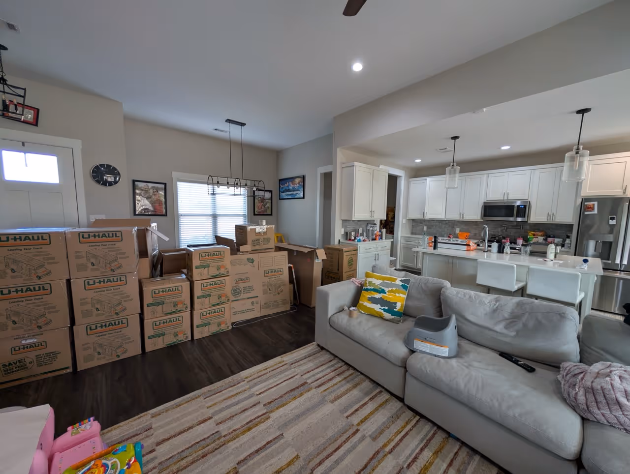 Living room and kitchen with packed U-Haul moving boxes stacked near a window, a gray sofa with colorful pillows, and kitchen island with white chairs.