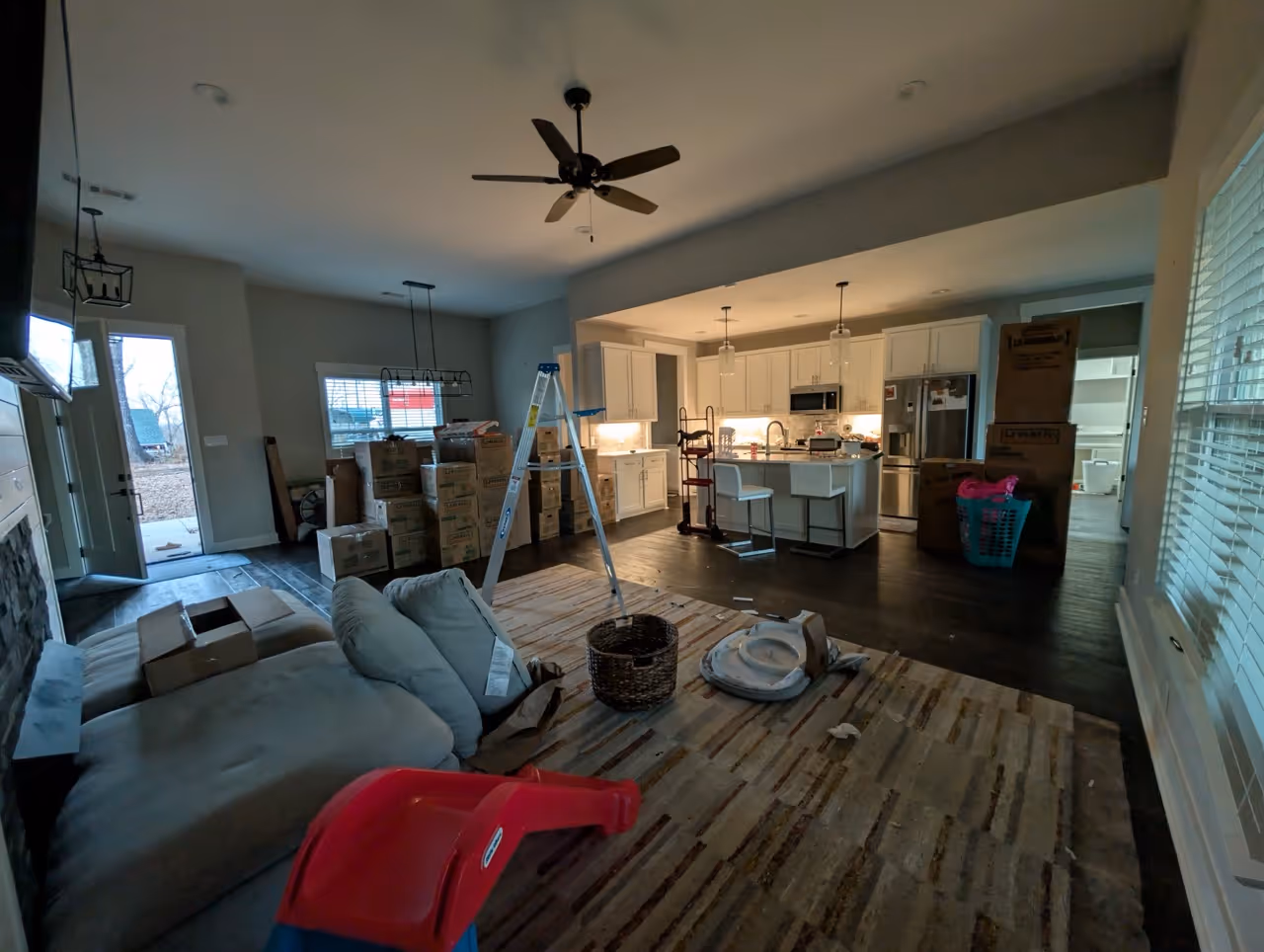 Open kitchen and living room area with unpacked moving boxes, a ladder, and furniture on a patterned rug.