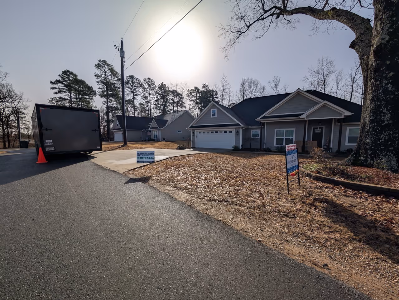 Suburban street view with a light gray house for sale, a large tree, a parked trailer, and two advertising signs on the lawn.