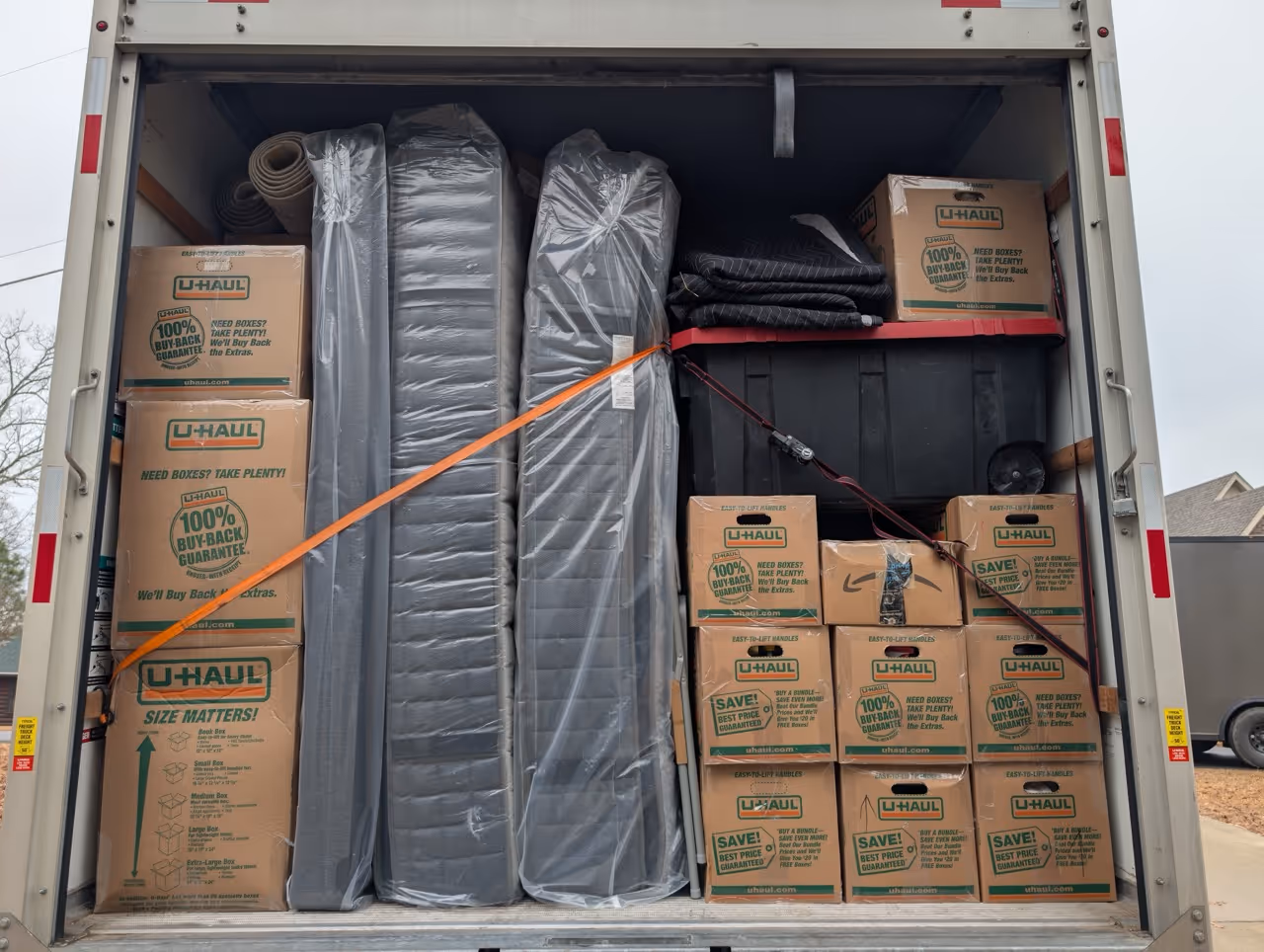 Back of a moving truck loaded with stacked U-Haul boxes, wrapped mattresses, rolled rugs, and a large plastic bin secured with orange and black straps.