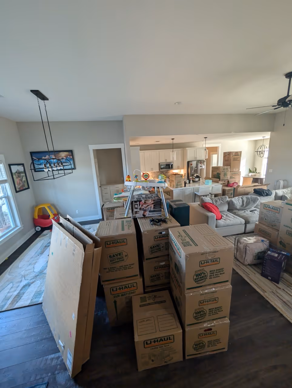 Living room and kitchen area filled with stacked U-Haul moving boxes, a red cushion on a gray sectional sofa, and a child's toy car near a window.
