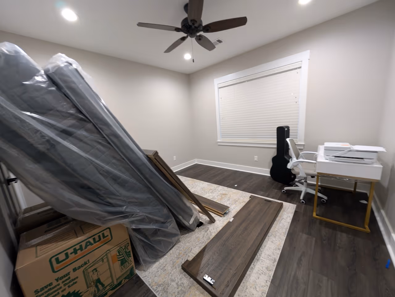 Room with light gray walls and dark wood floor, containing packed furniture parts, a U-Haul box, a white office chair, a black guitar case, and a printer on a small desk.
