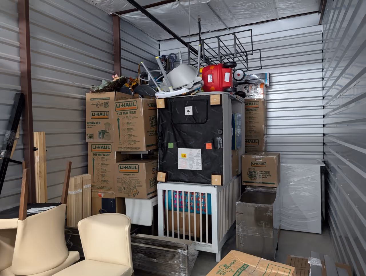 Storage unit filled with stacked U-Haul boxes, beige chairs, a white crib, and various household items including a red toy car and a black chair upside down.