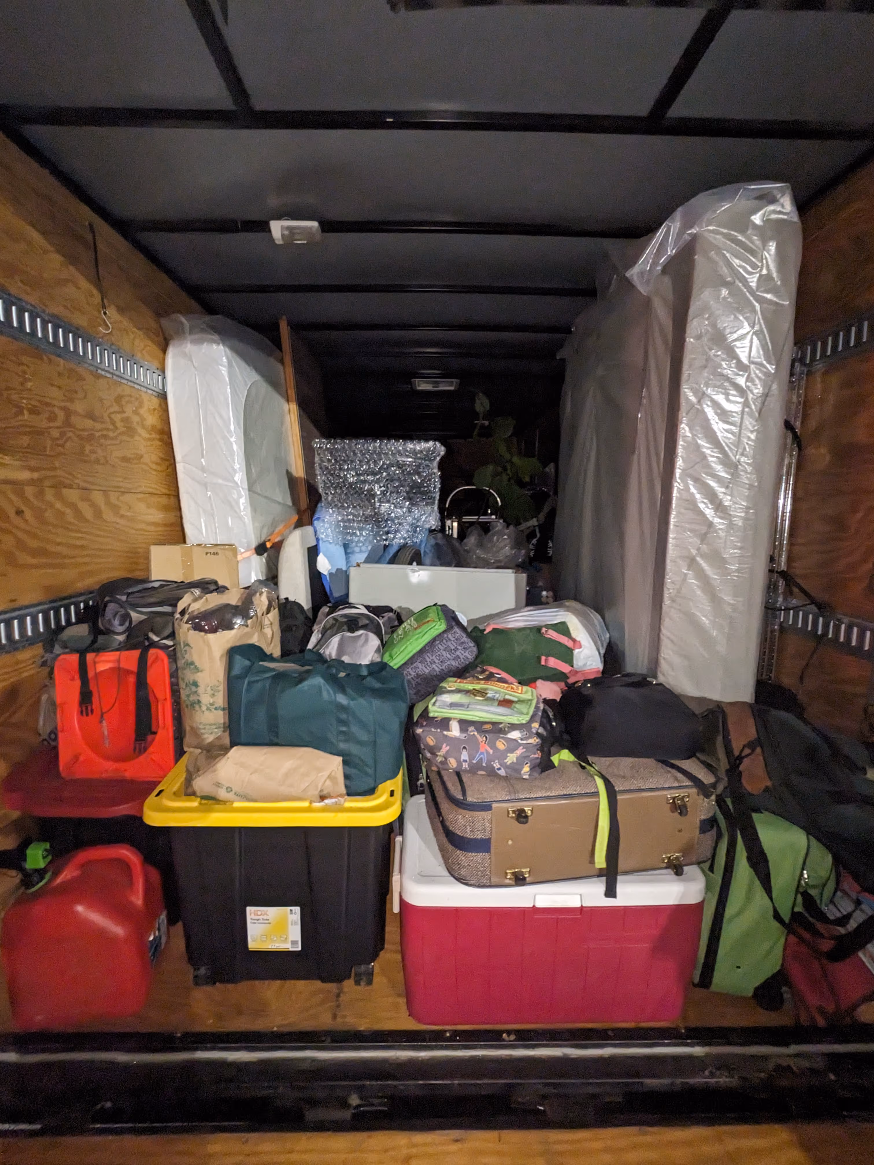 Interior of a moving truck loaded with packed items including mattresses, suitcases, coolers, bags, and a red gas can.