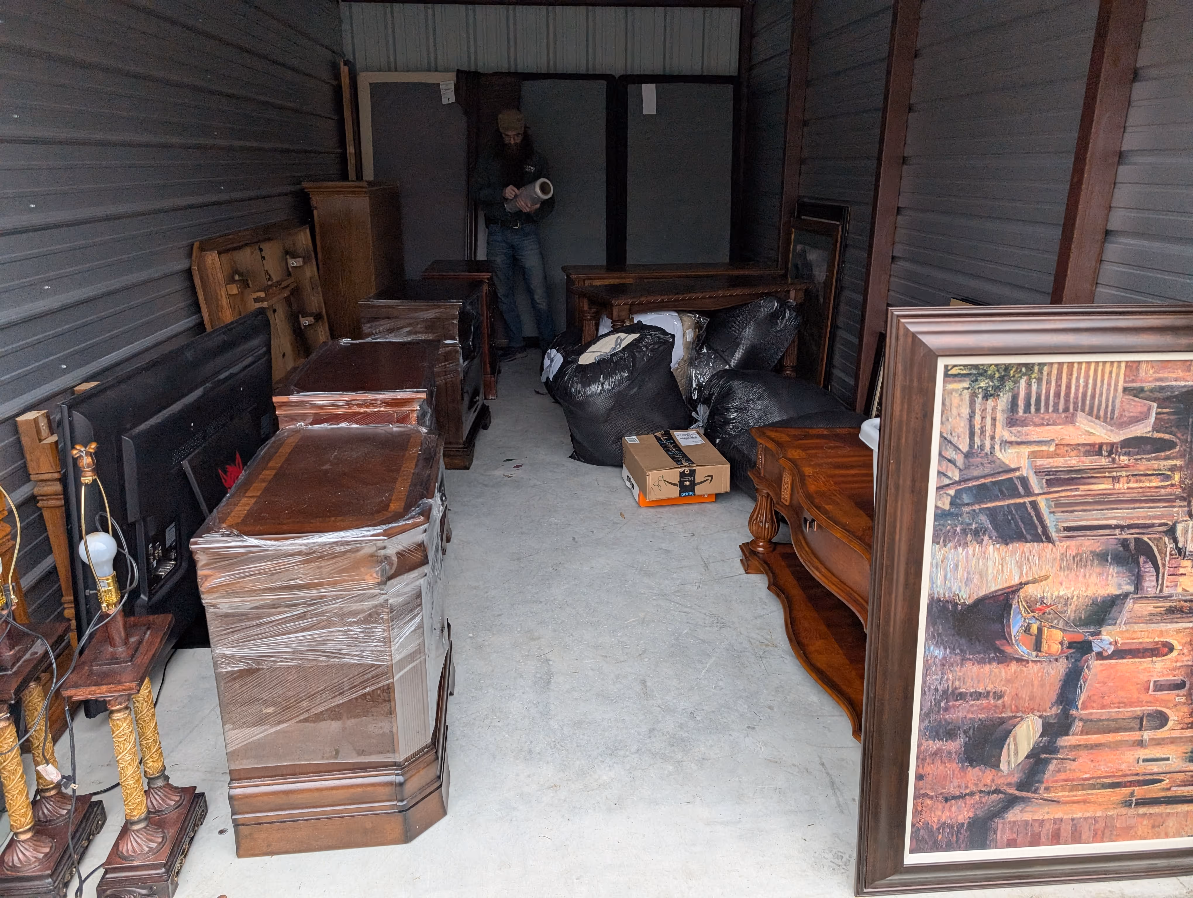 Storage unit filled with wooden furniture wrapped in plastic, black trash bags, a large framed painting, and a person standing in the back holding an object.