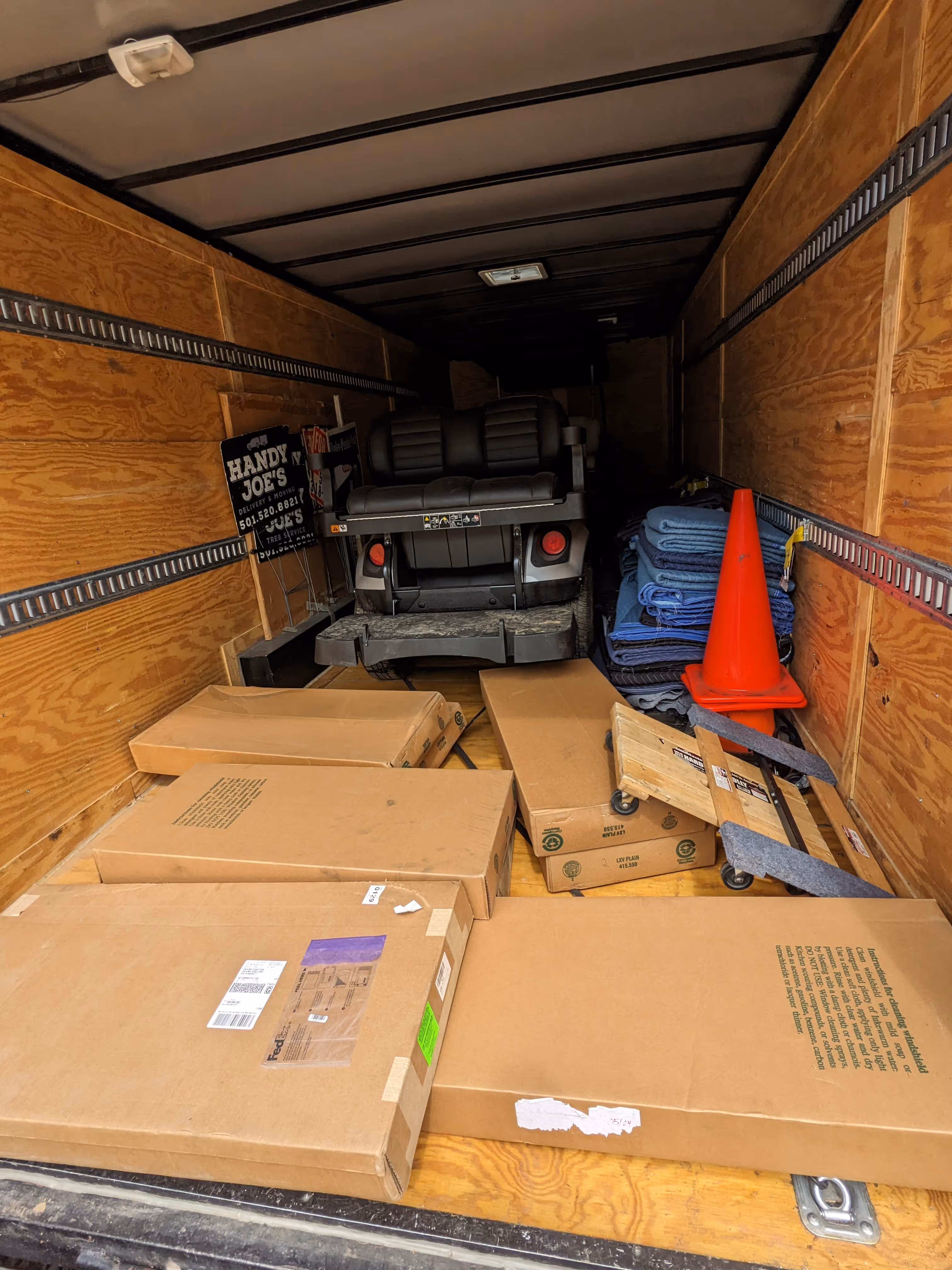 Inside a wooden moving truck with stacked cardboard boxes, a dolly, moving blankets, an orange traffic cone, and the back of a golf cart.