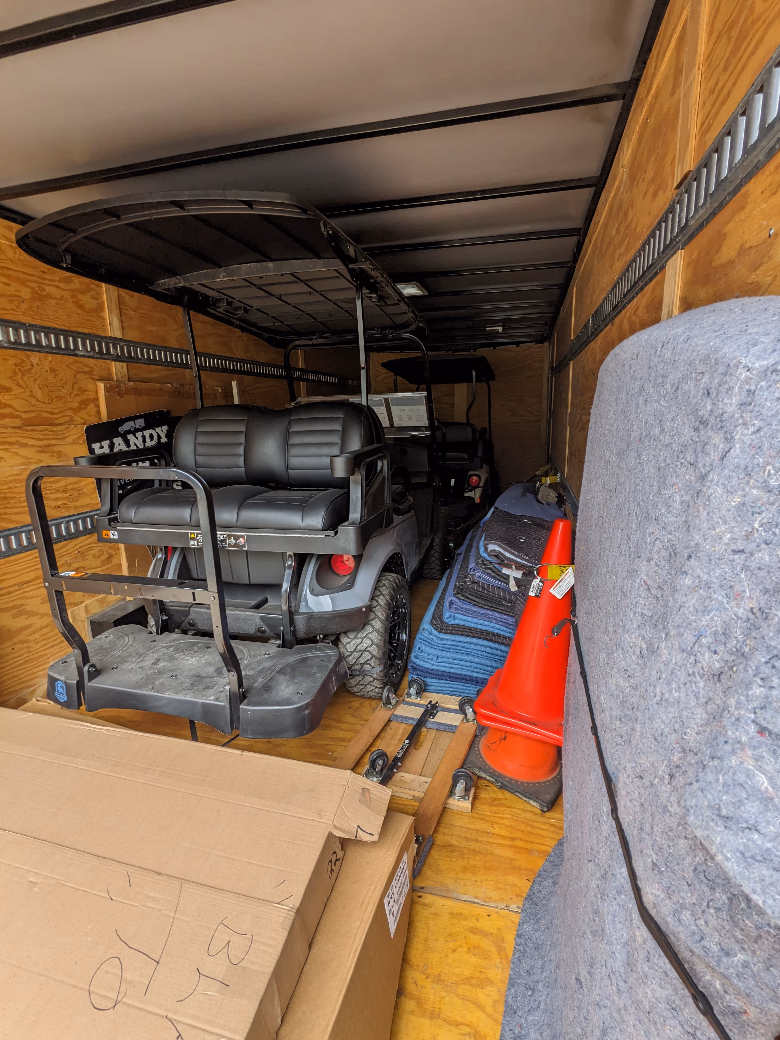 Interior of a trailer with two black golf carts, orange traffic cones, stacked moving blankets, and a large cardboard box.
