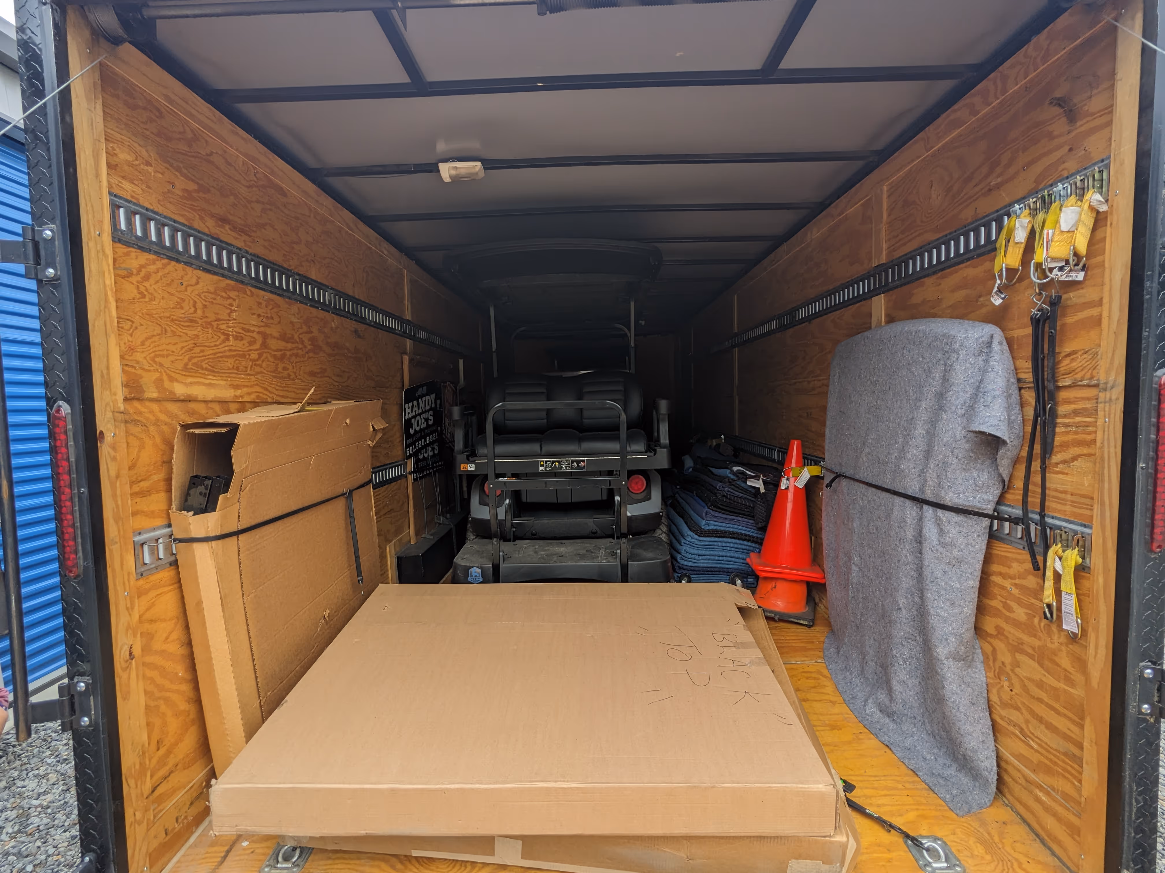 Interior of a wooden trailer loaded with cardboard boxes, a golf cart, stacked moving blankets, orange traffic cones, and straps securing the items.