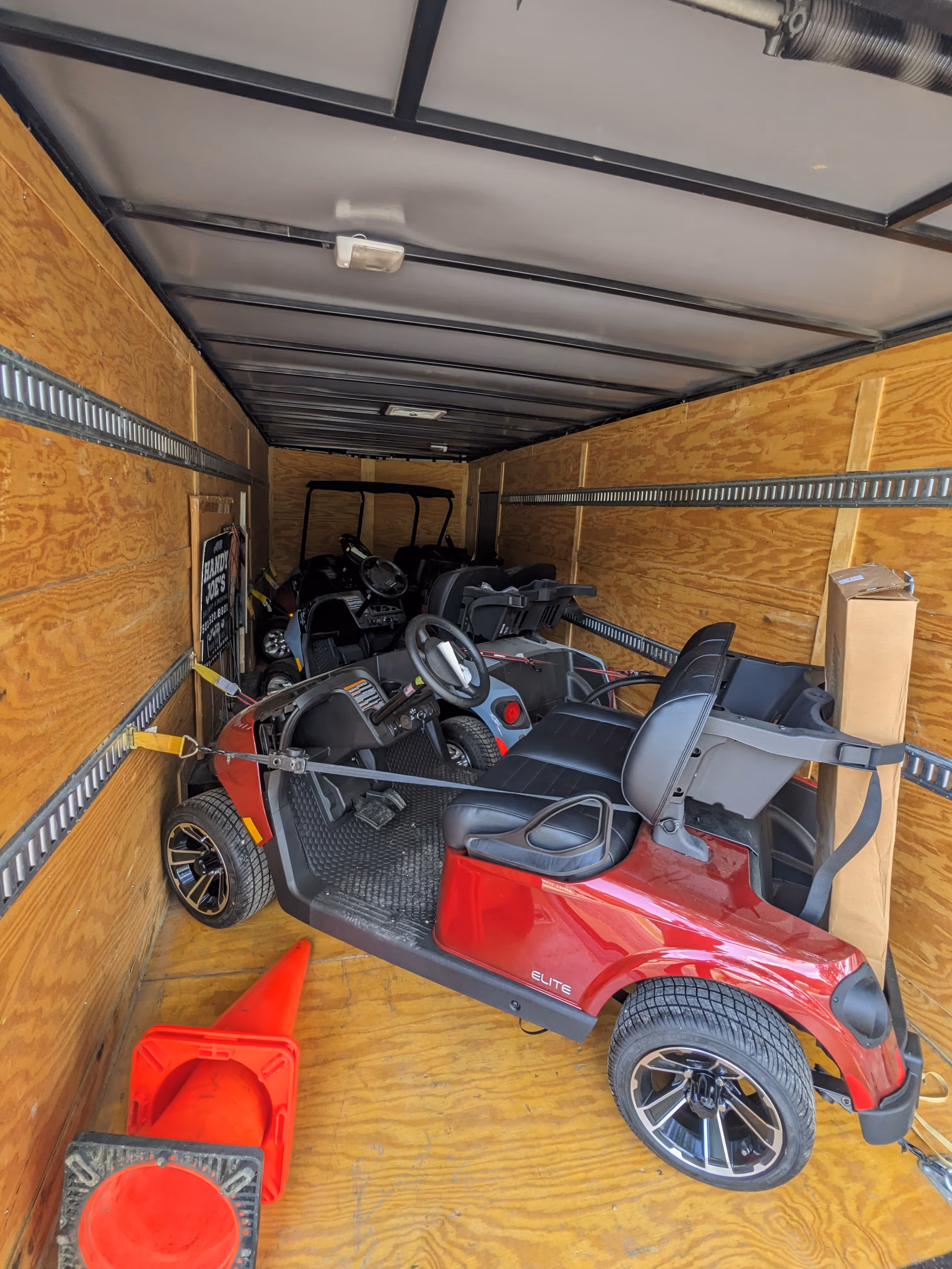 Red golf cart secured inside a wooden trailer with several other golf carts and two orange traffic cones on the floor.