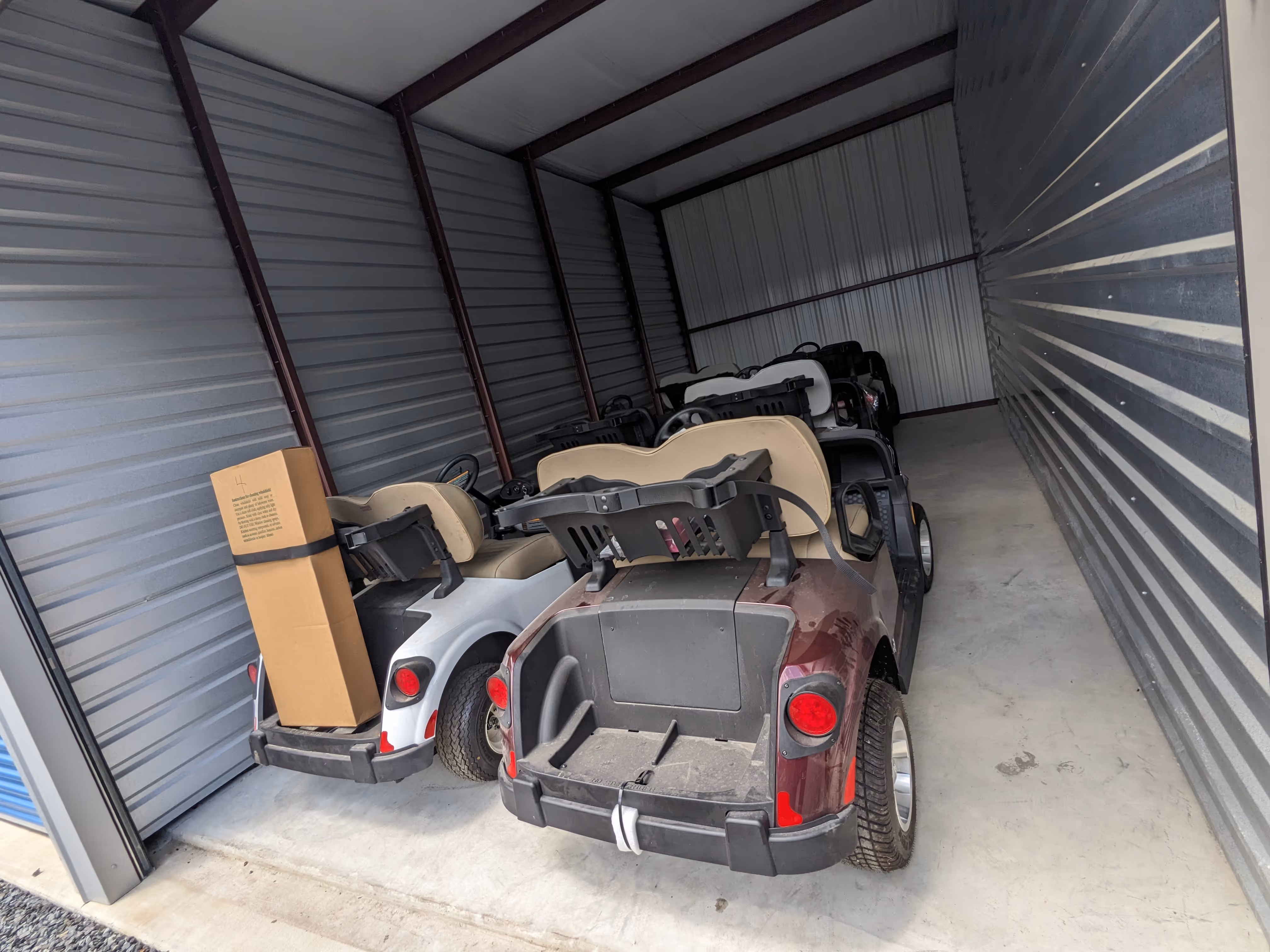 Several golf carts parked inside a metal storage unit, with one white cart holding a tall cardboard box strapped at the back.