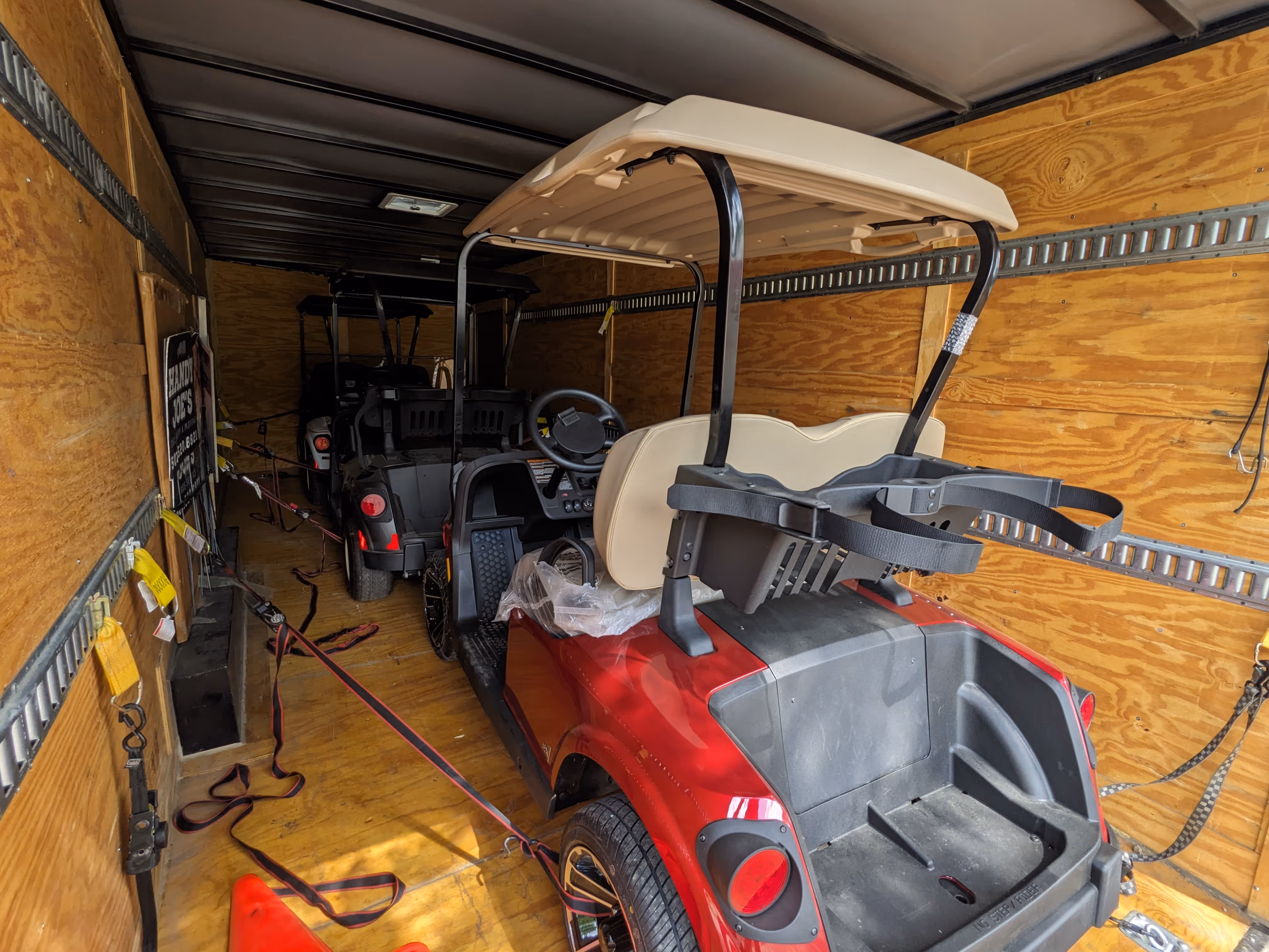 Two golf carts, one red and one black, secured inside a wooden trailer with tie-down straps.