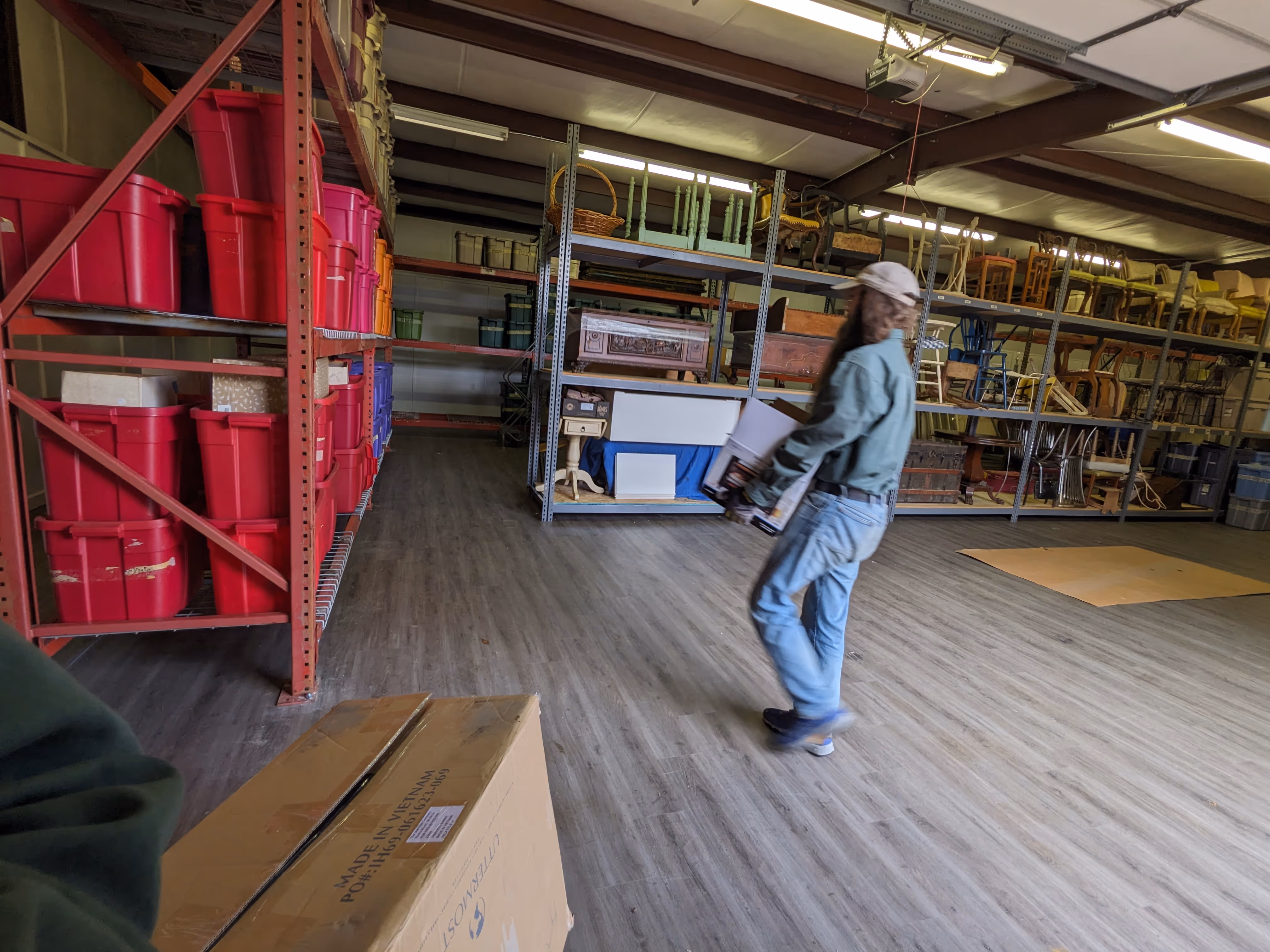 A person wearing a green jacket and cap carrying a box in a storage room with shelves filled with red bins and assorted chairs.
