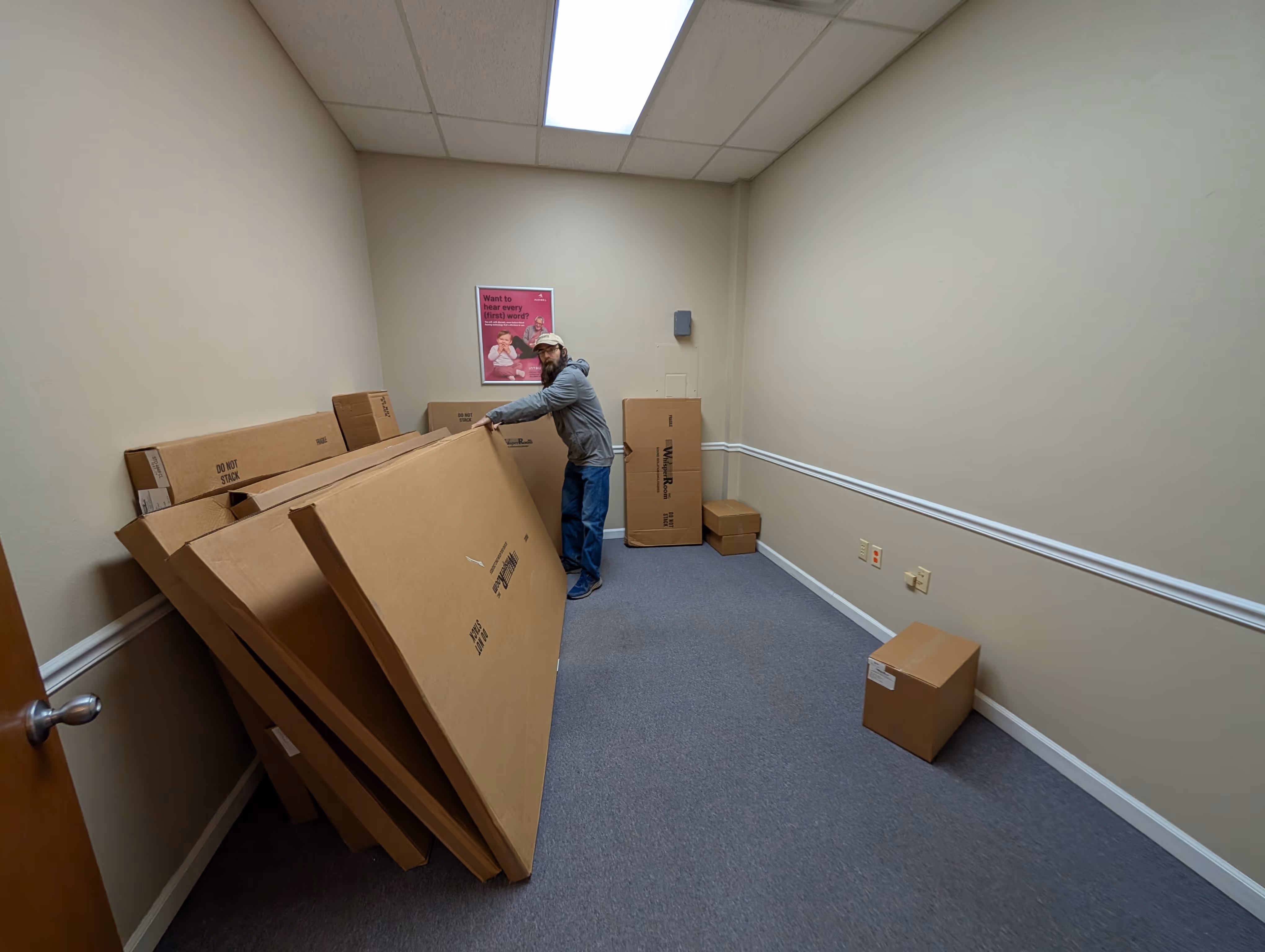 Man in a cap and glasses organizing large flat cardboard boxes in a small beige-walled room with carpeted floor.