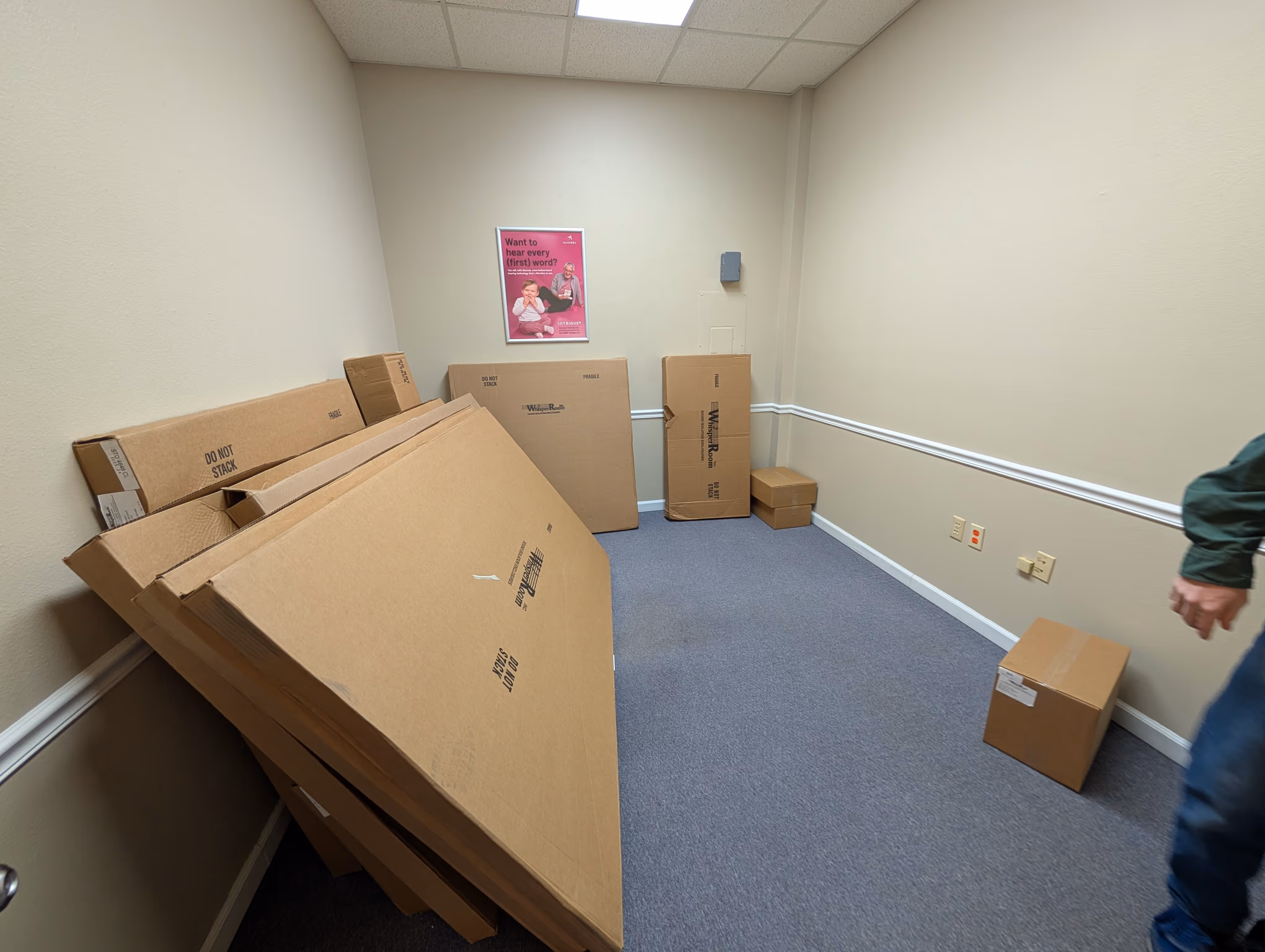 Small empty office room with beige walls and blue carpet, containing stacked large cardboard boxes leaning against one wall and smaller boxes on the floor.