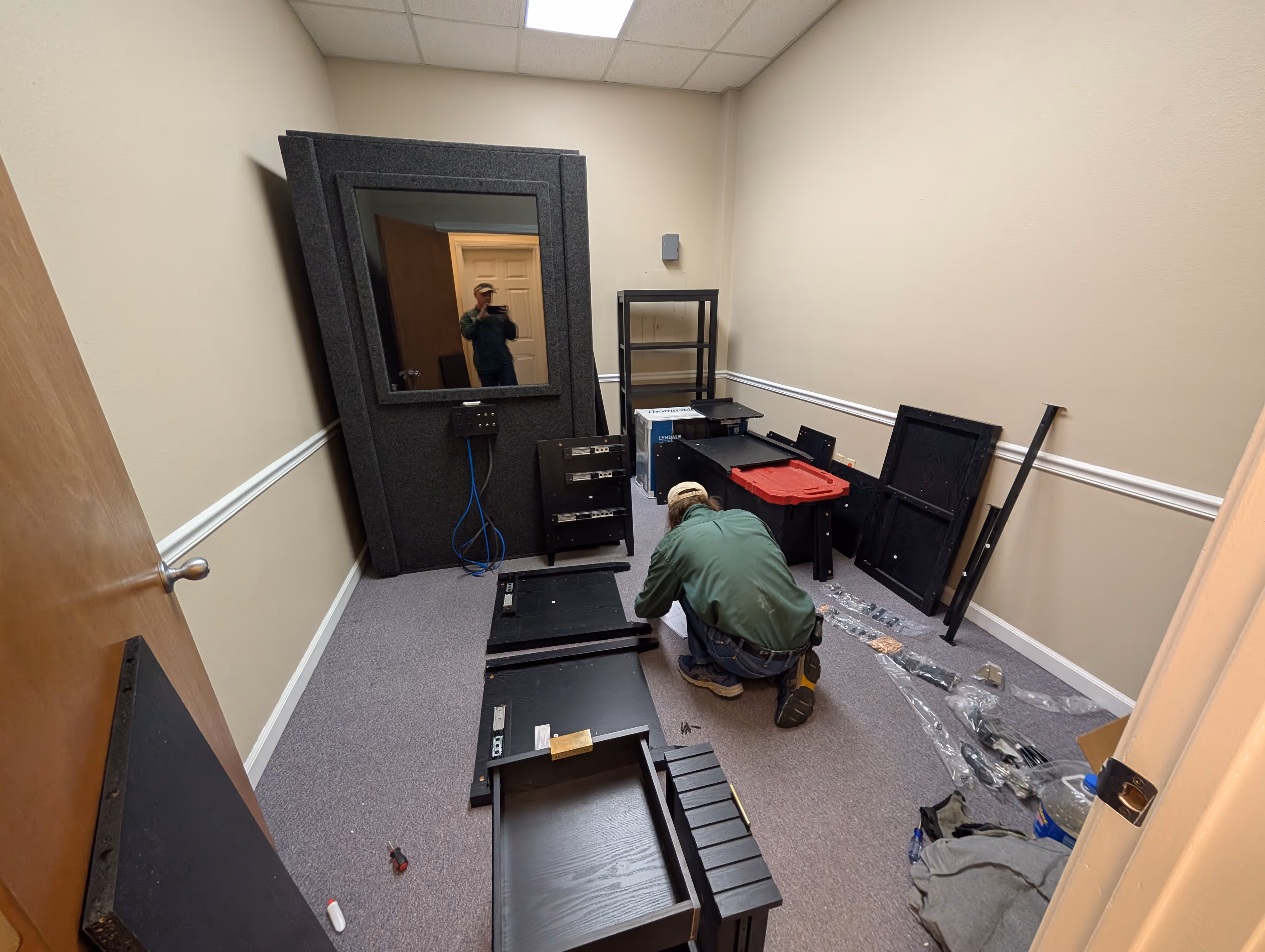 Man assembling black wooden furniture pieces spread out on the floor inside a beige-walled room with a mirror and shelves.