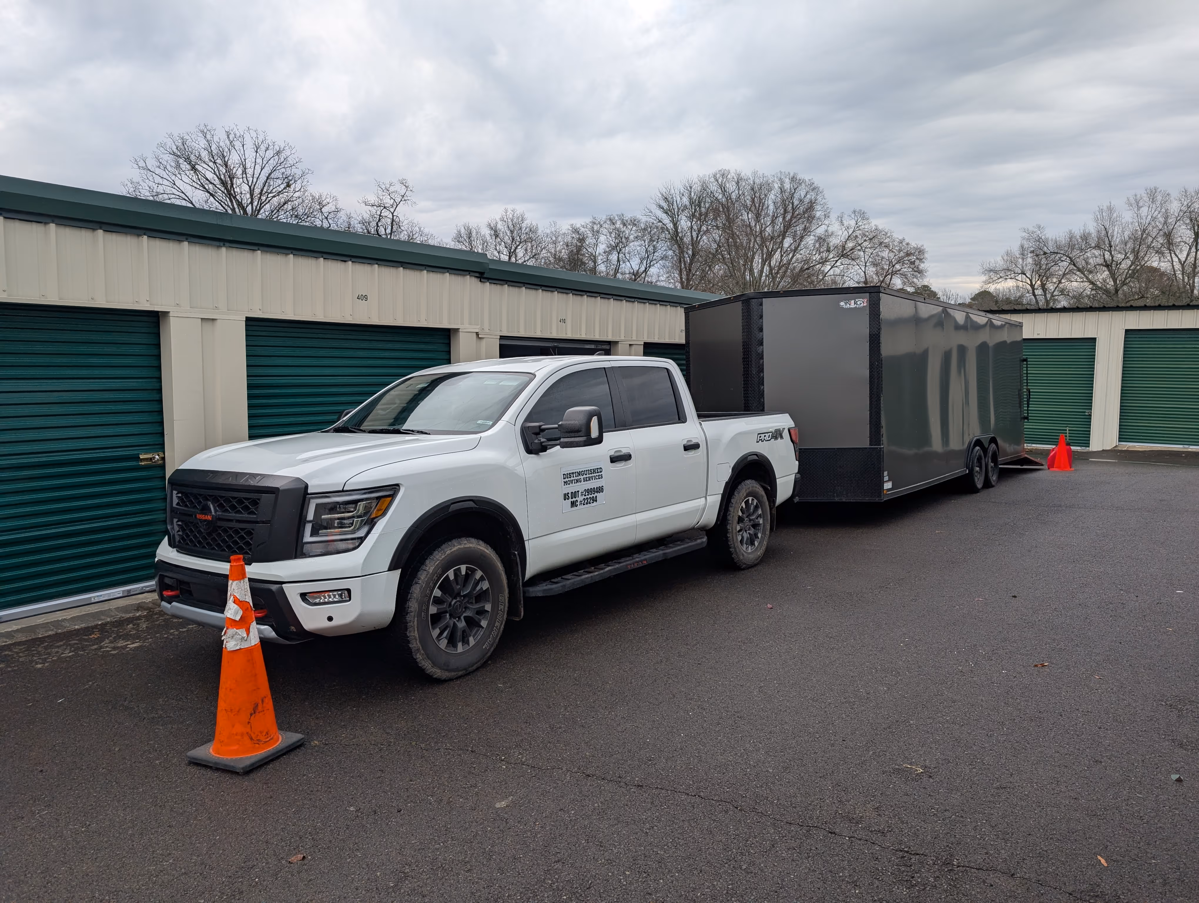 White Nissan pickup truck with company decal parked in front of green storage unit doors, towing a large black enclosed trailer with orange cones placed near the truck and trailer.